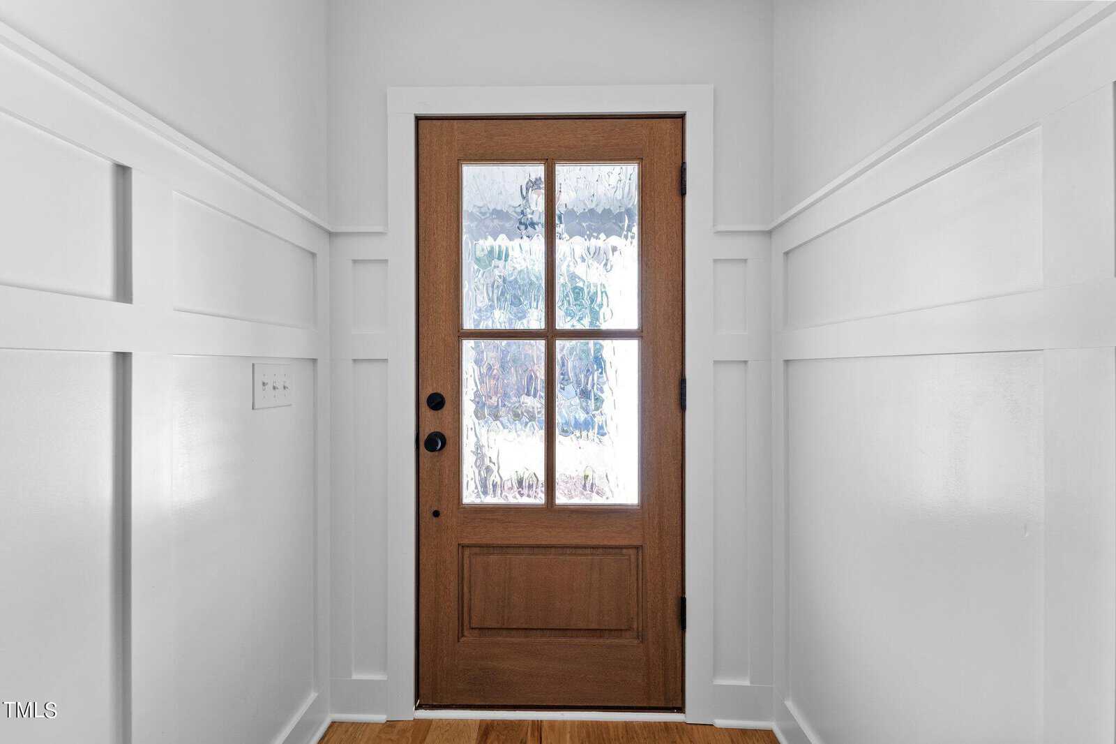 A wooden door with glass panels stands at the end of a narrow hallway with white paneled walls. Sunlight casts soft shadows through the textured glass of the door.