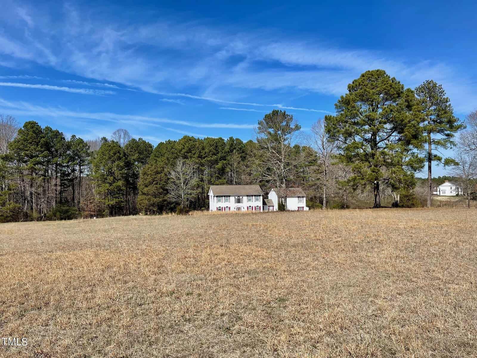A rural landscape featuring a wide, open field of dried grass under a clear blue sky, with a white house and several trees in the background.