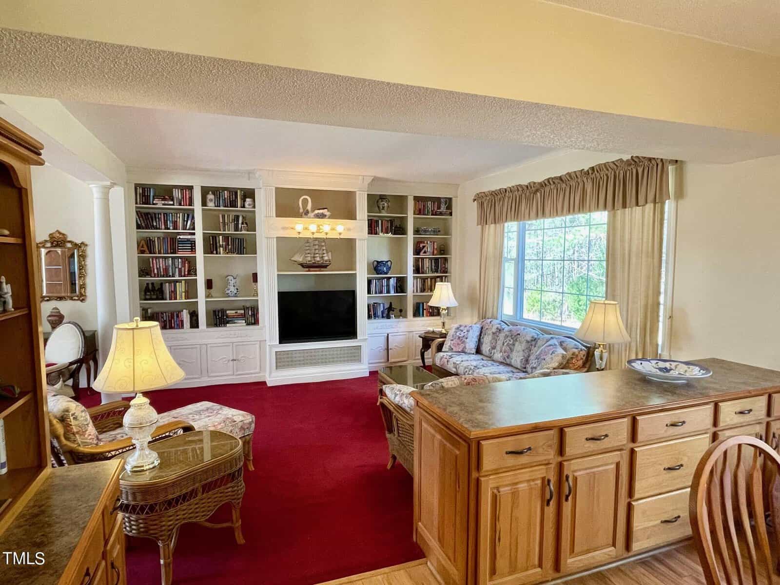 A cozy living room featuring a red carpet, white built-in bookshelves filled with books, two table lamps, a floral sofa, and a wooden secretary desk.