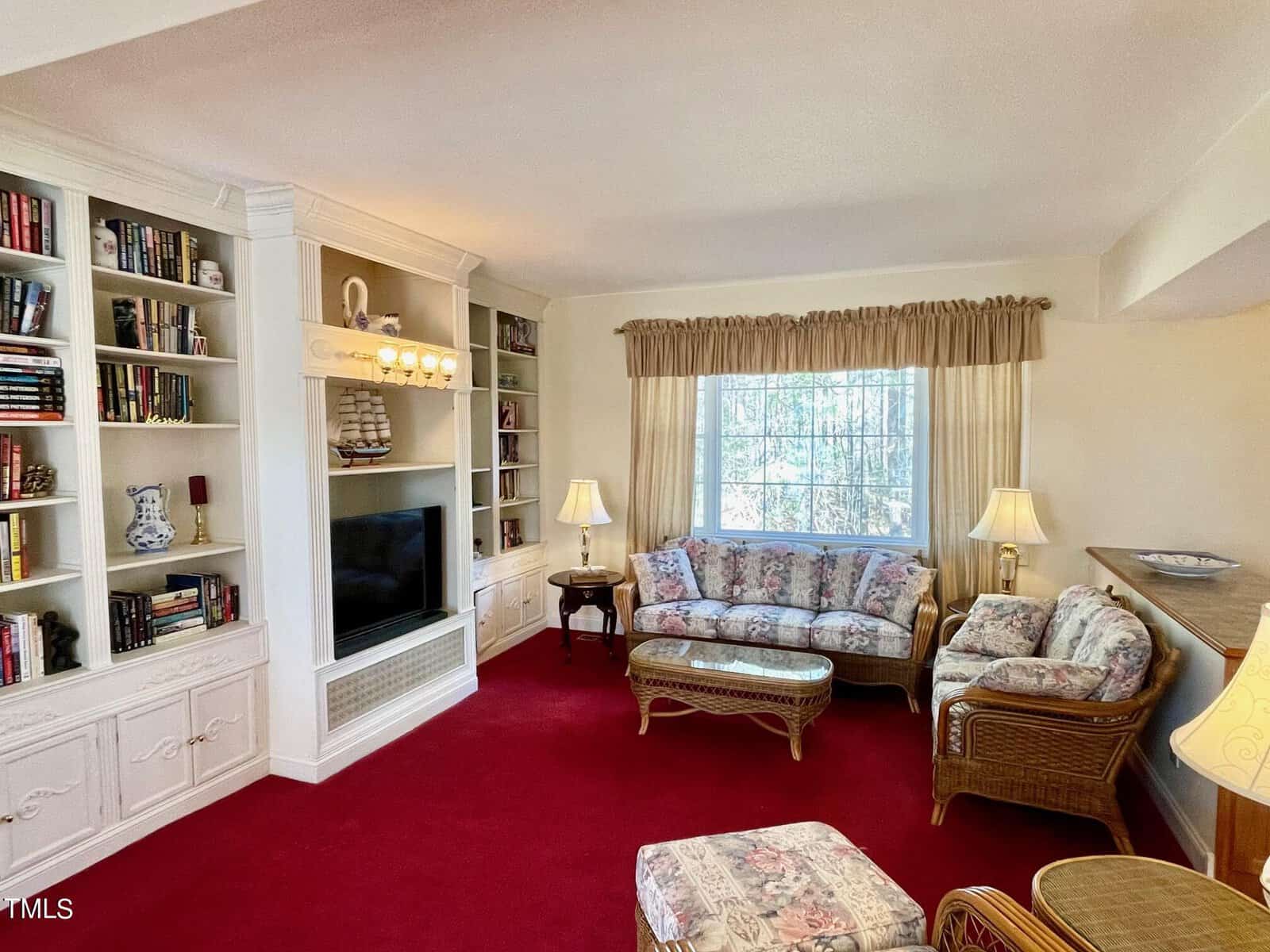 A cozy living room featuring a white built-in media cabinet, floral sofas, and a red carpet. natural light streams in through a large window, with a traditional decor style.