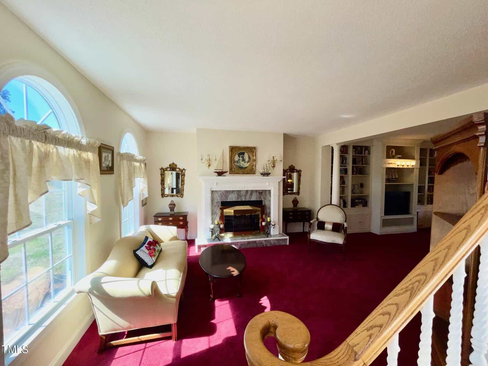 Elegant living room with a white sofa, a fireplace, red carpet, and large windows with curtains. stairs with wooden railing are visible on the right.