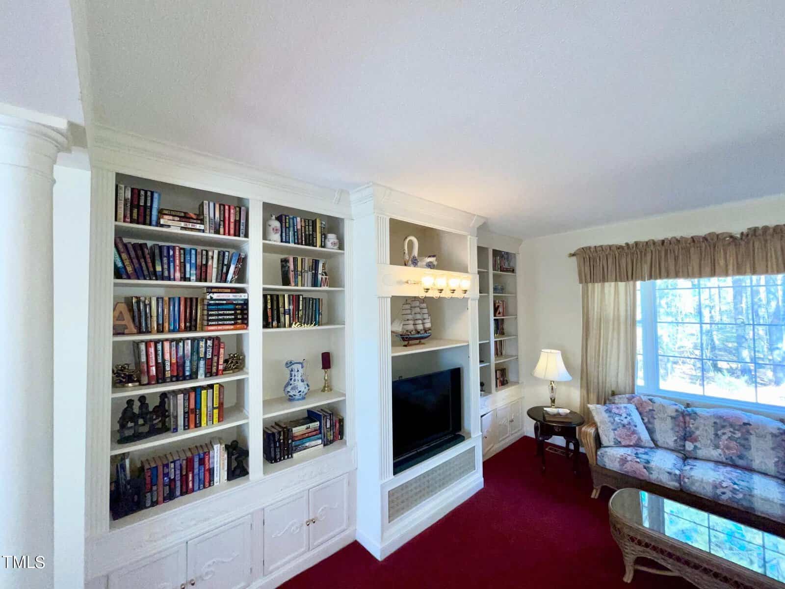 A cozy living room with white built-in bookshelves filled with books, a white fireplace, and a tv. the room has a red carpet, a floral sofa, a small table, and a curtained window with natural light.