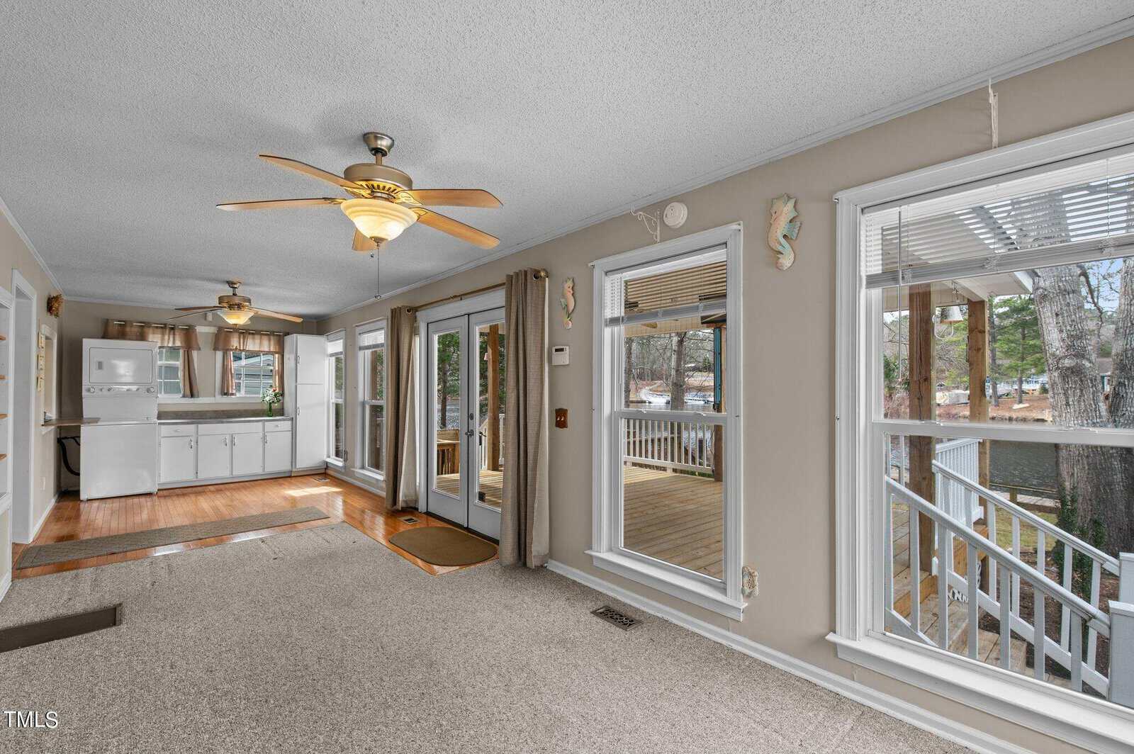 A bright sunroom with beige walls, carpeted floors, ceiling fans, large windows, and glass doors leading to a porch. The kitchen area with white cabinets and appliances is visible on the left.