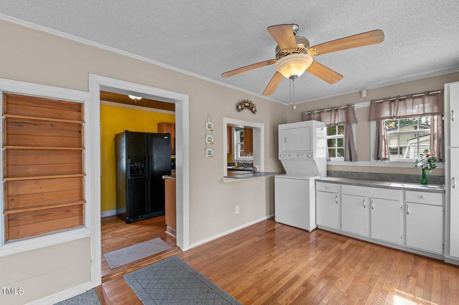 Bright kitchen with hardwood floors, white cabinets, stacked washer and dryer, a ceiling fan, small window, and a black refrigerator in an adjacent yellow room. A vase with flowers sits on the counter by the window.