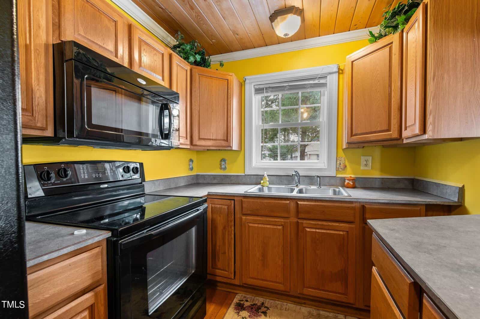 A small kitchen with wooden cabinets, black appliances, yellow walls, a central window above the sink, and gray countertops. Plants sit on top of the cabinets and a light fixture is on the wooden ceiling.