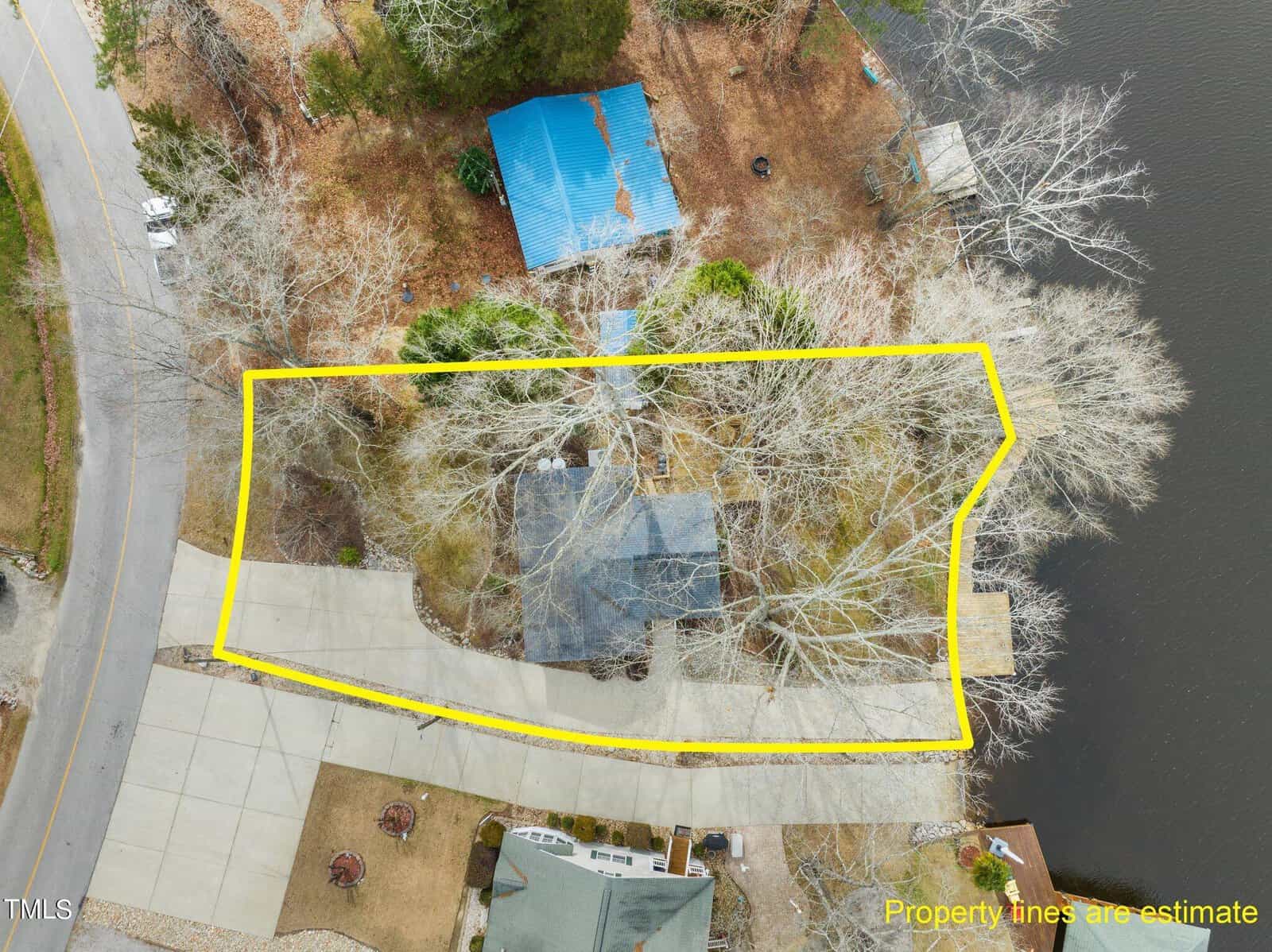 Aerial view of a house and yard outlined in yellow, located between a road and a body of water, surrounded by trees and neighboring properties. The property lines are labeled as estimates.