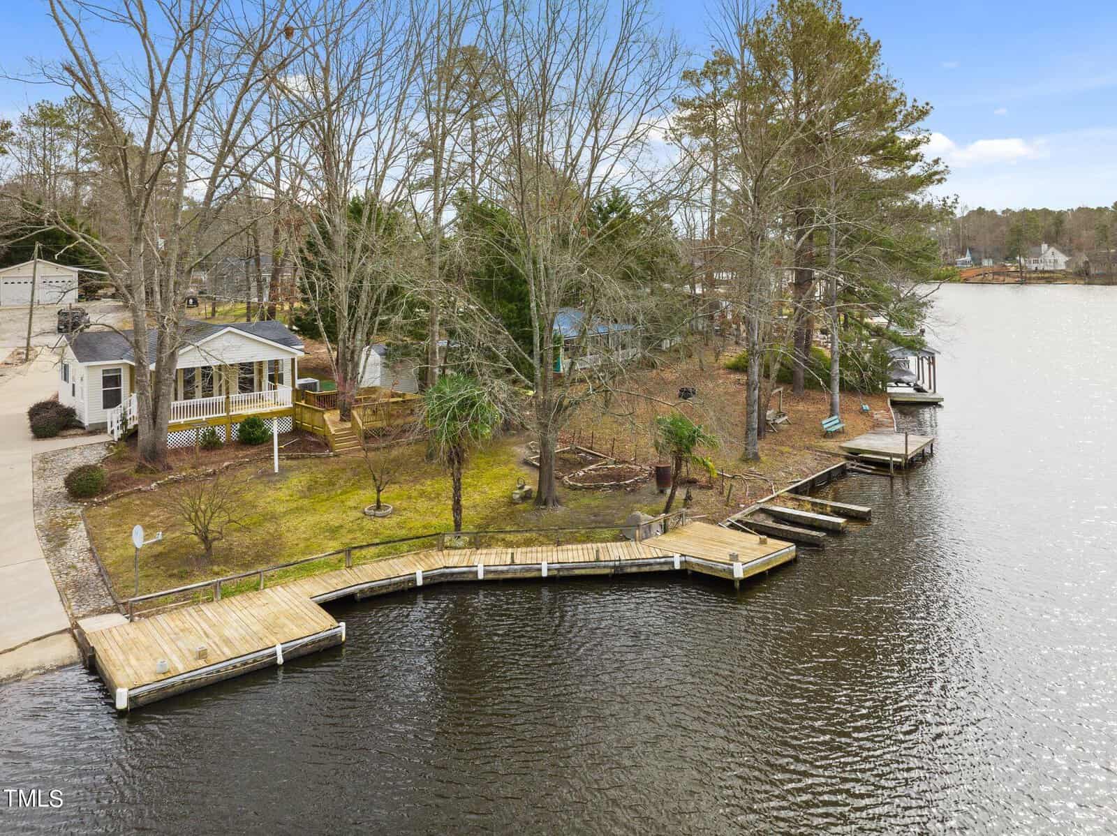 A lakeside property with a small house, several trees, and a wooden dock extending along the shoreline. The yard is grassy with landscaping, and the water is calm under a partly cloudy sky.