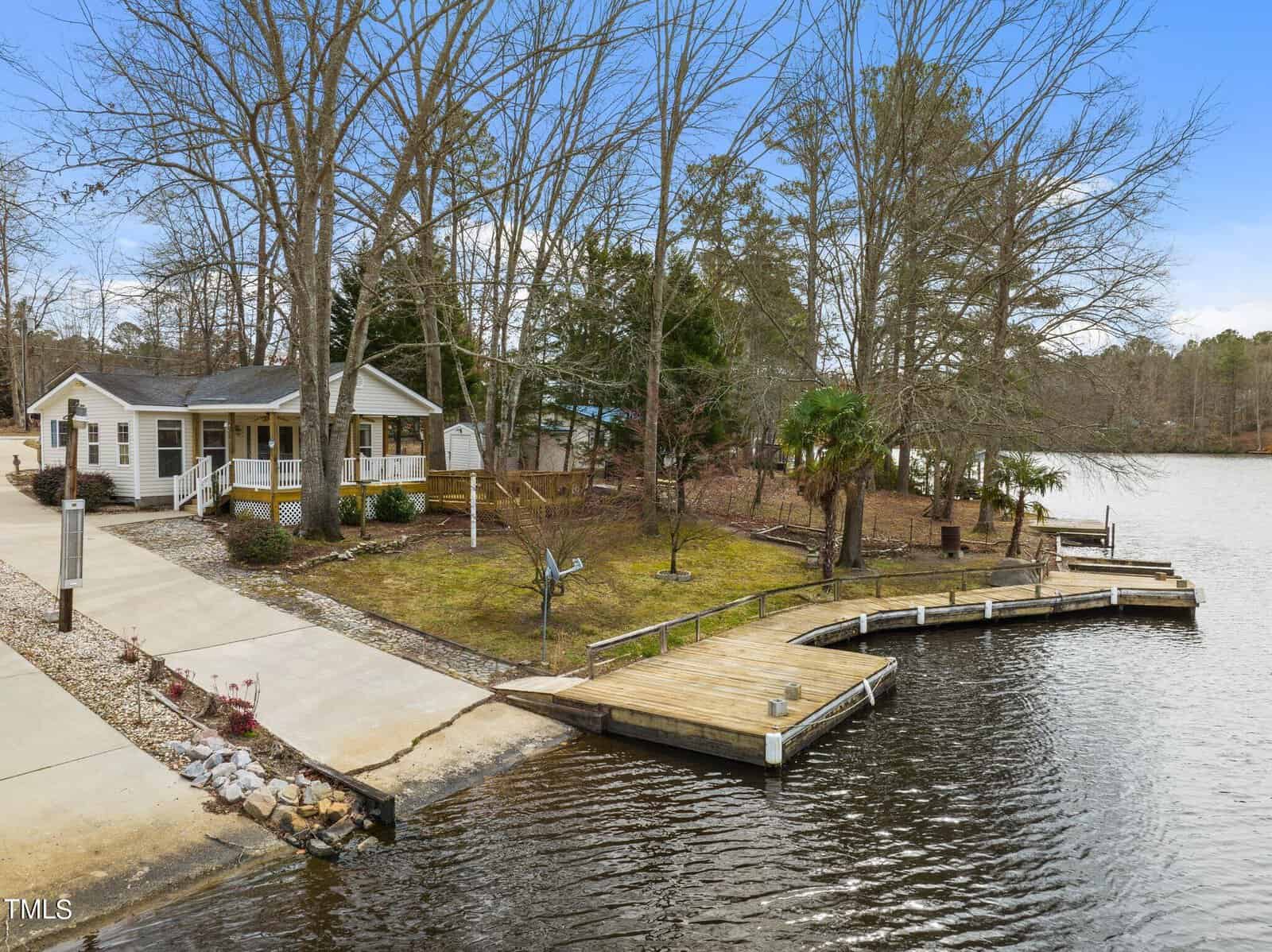 A small house with a porch sits near a wooded shoreline, next to a curved wooden dock extending into a calm lake. Leafless trees surround the property, and the sky is partly cloudy.