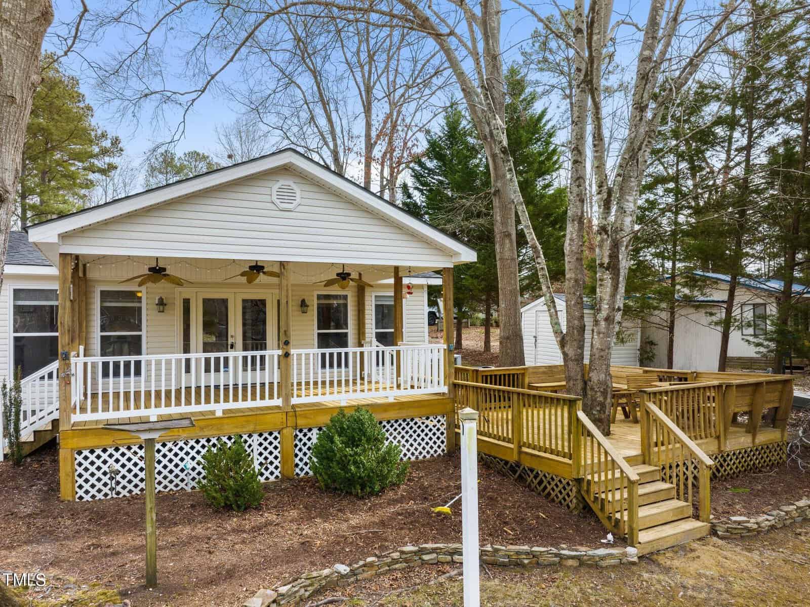 A cozy white house with a large wooden front porch and deck, surrounded by trees. The porch has ceiling fans, railings, and steps leading down to the yard with some shrubs and garden beds.