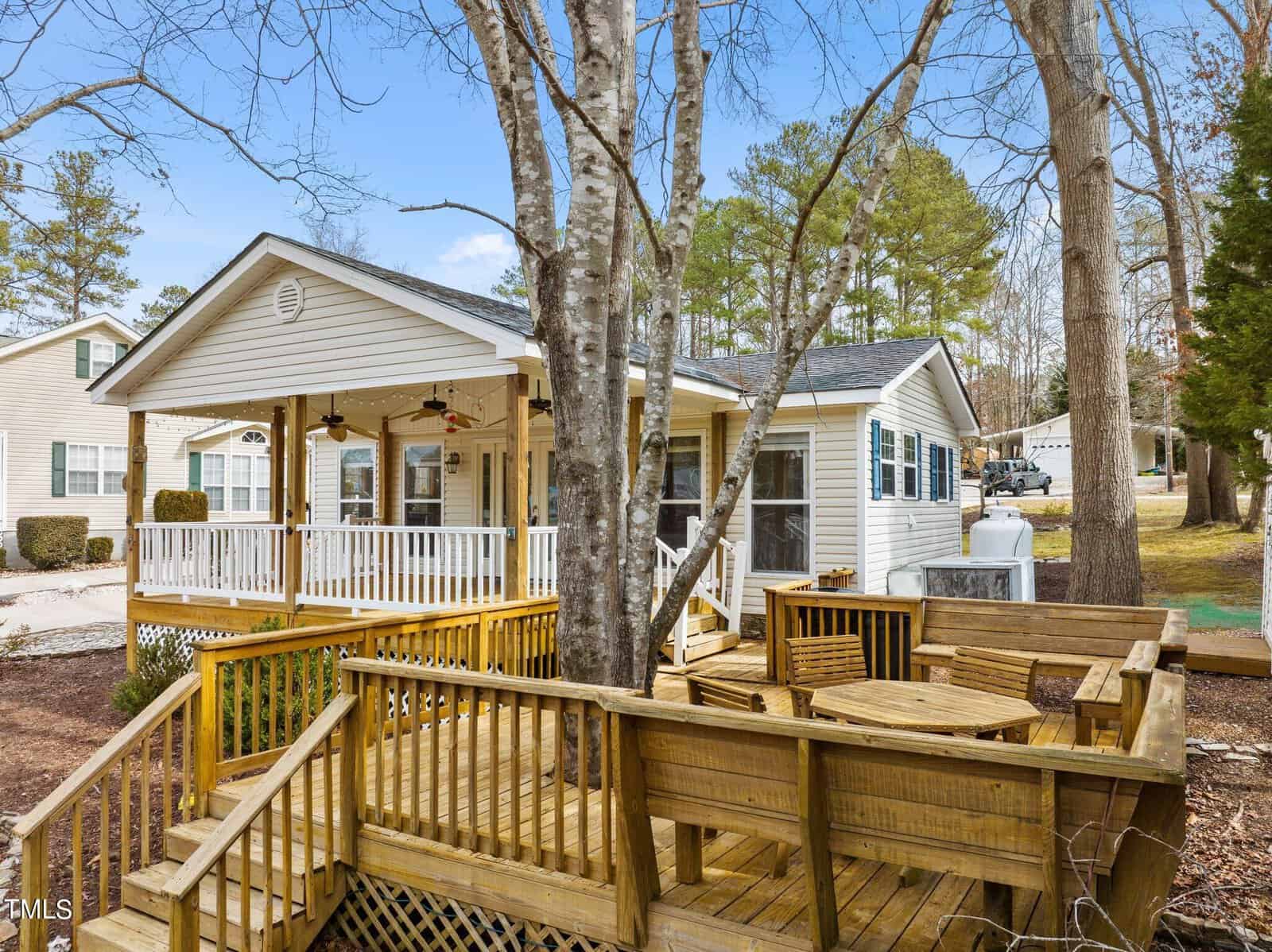 Single-story house with a large white porch, surrounded by trees, and a wooden deck area with built-in seating in the foreground. The yard is landscaped with mulch and the sky is clear.