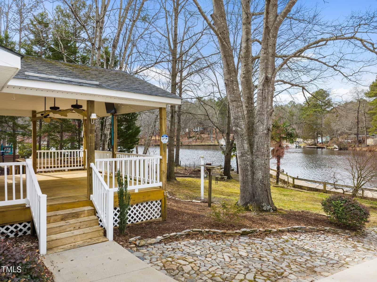 A wooden deck with white railings overlooks a peaceful lake surrounded by trees. A stone pathway leads from the deck toward the water, and houses are visible across the lake under a cloudy sky.