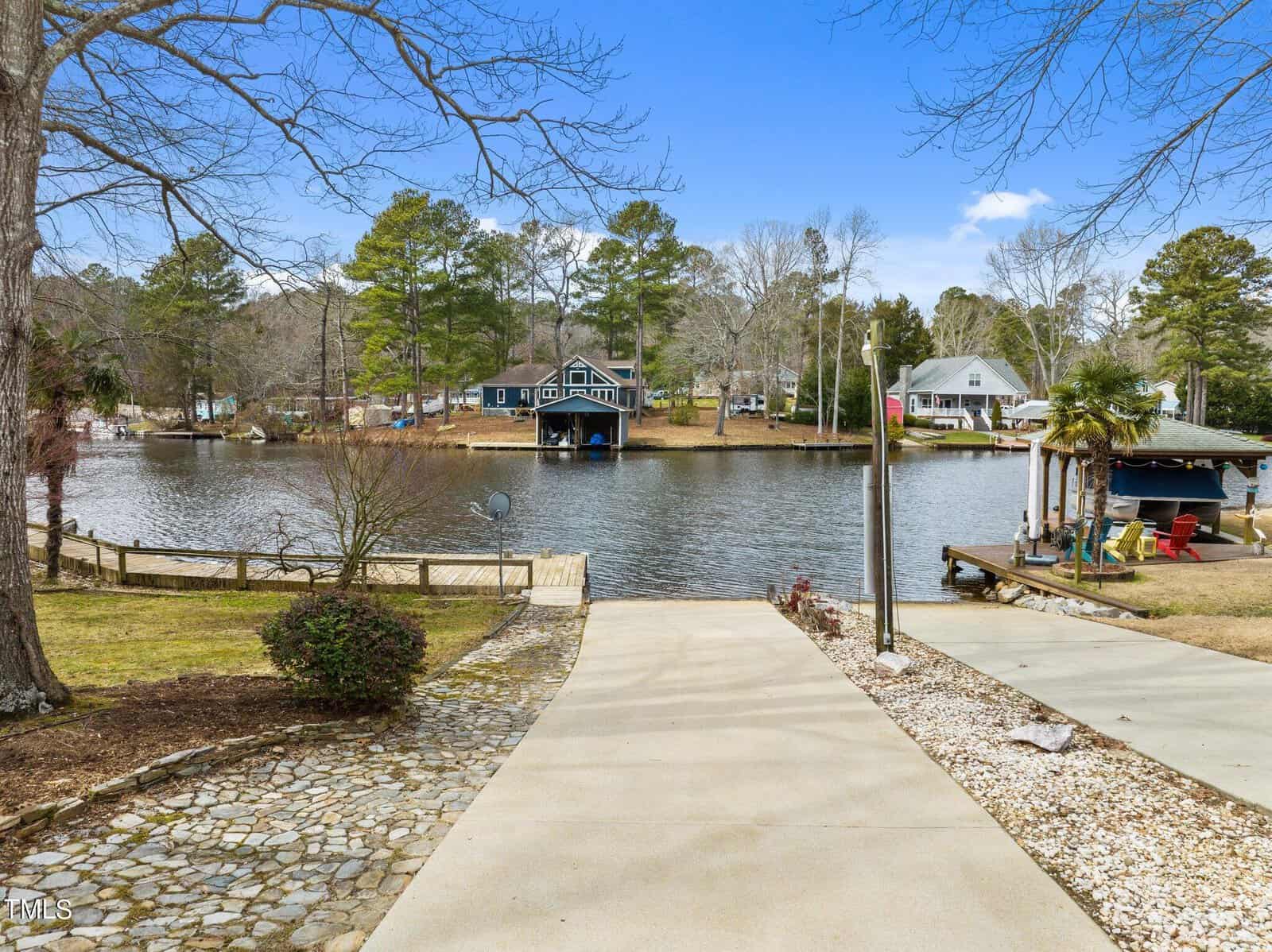 A paved driveway leads to a lakeside dock, with a calm lake, a boathouse, and trees visible across the water under a clear blue sky.
