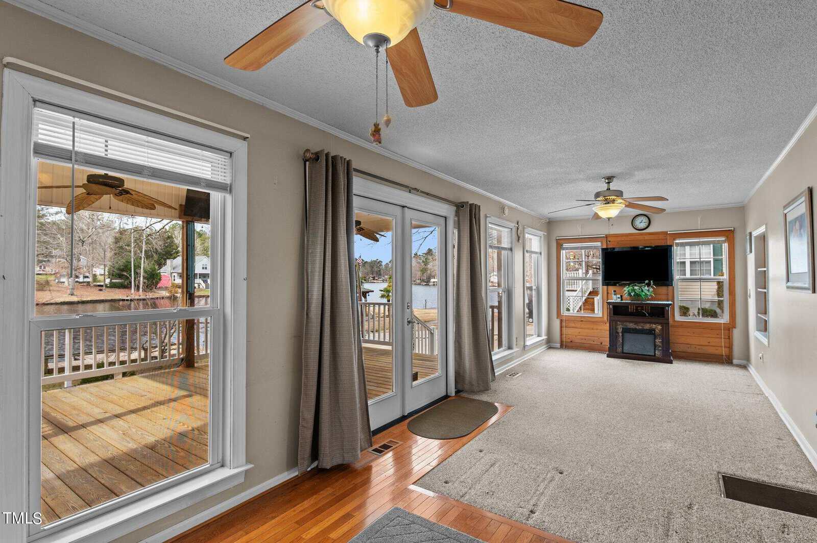 A bright sunroom with large windows, double glass doors leading to a wooden deck, and two ceiling fans. The room has beige walls, carpet, hardwood flooring, and a TV on a wooden stand. A lake is visible outside.