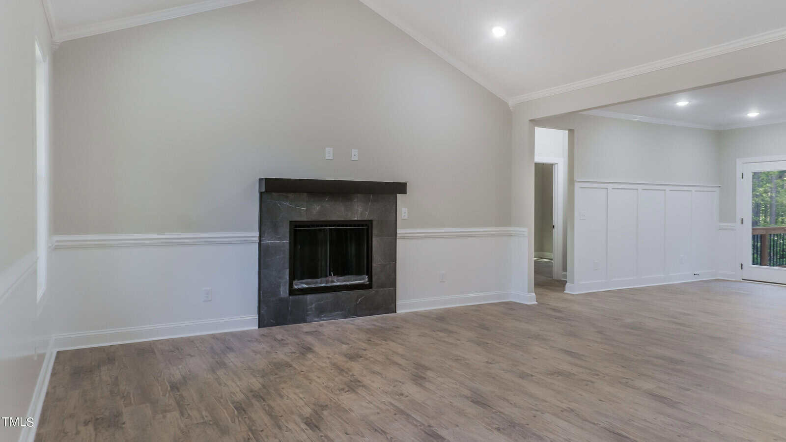 A spacious, empty room with light-colored walls and wooden flooring features a black fireplace centered on the left wall. The ceiling is vaulted, and recessed lighting is present. There's an open doorway leading to another area, and a glass door opens to a deck outside.