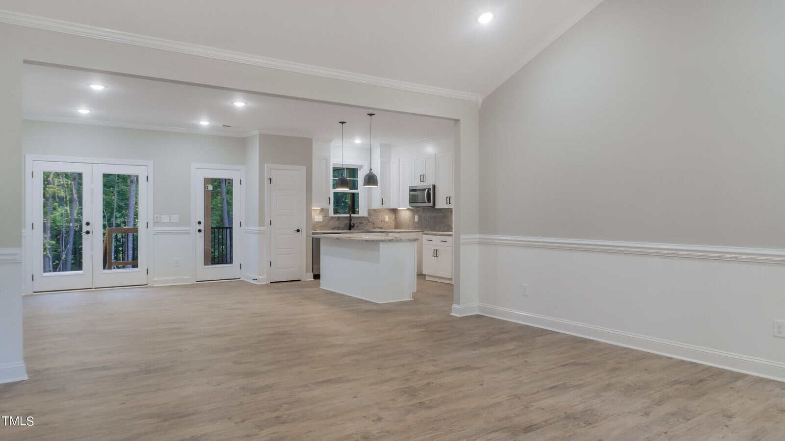 An open-concept living area with a view into a modern kitchen. The living space has light-colored wood flooring, and French doors lead to an outdoor space. The kitchen features a central island, pendant lighting, white cabinets, and stainless steel appliances.
