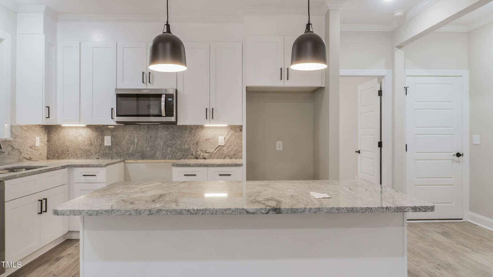 Modern kitchen with white cabinetry and a large granite island countertop. Two black pendant lights hang over the island. The backsplash features a matching granite design. A stainless steel microwave is mounted above the countertop. White walls and a wooden floor complete the look.