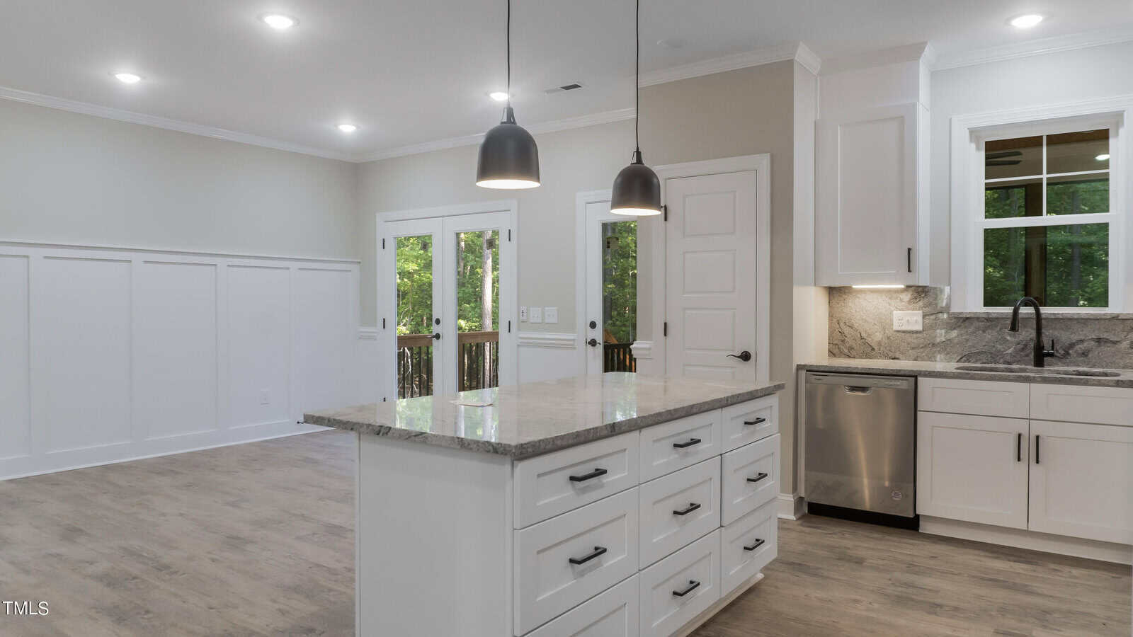 A modern kitchen features white cabinetry, a large island with a marble countertop, pendant lighting, and stainless steel appliances. The adjacent dining area has hardwood floors, wainscoting, and glass doors leading to a patio, with large windows providing ample natural light.