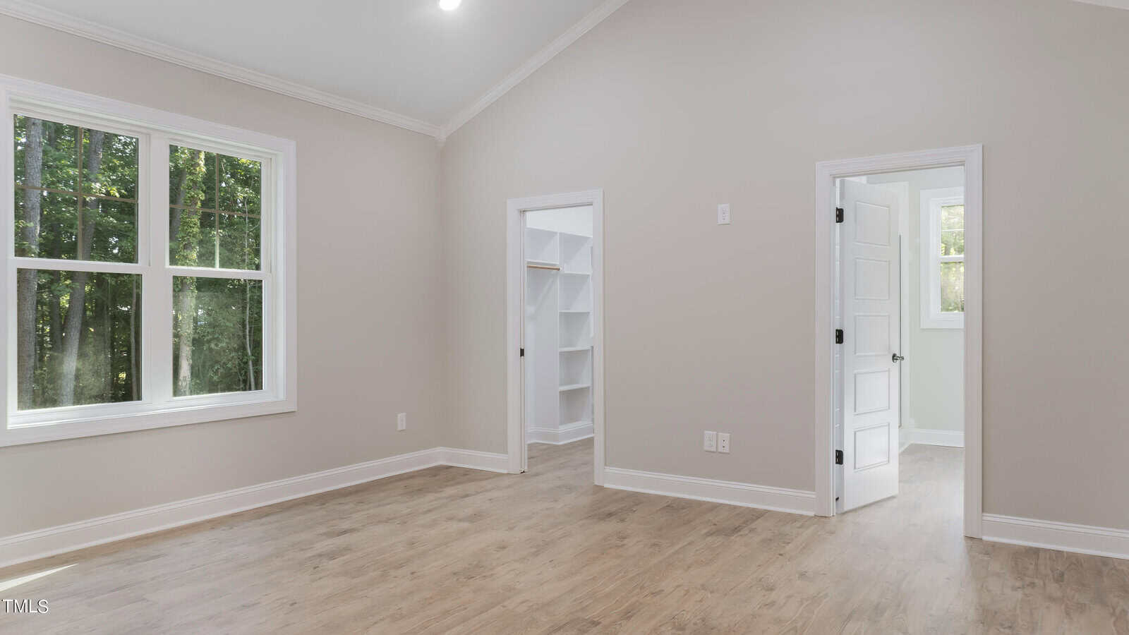 This image shows an empty, modern bedroom with light neutral walls, large windows on one side letting in natural light, a hardwood floor, and a white door leading to a closet with built-in shelves and another white door leading to another room.