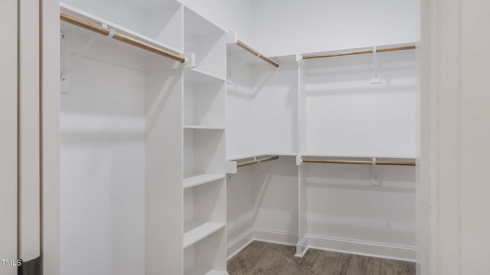 A walk-in closet with white walls, wooden hanging rods, and built-in shelves. The floor is covered with dark wood laminate, and the space is brightly lit by natural light. The closet offers ample storage for clothes and accessories with multiple hanging and shelving options.