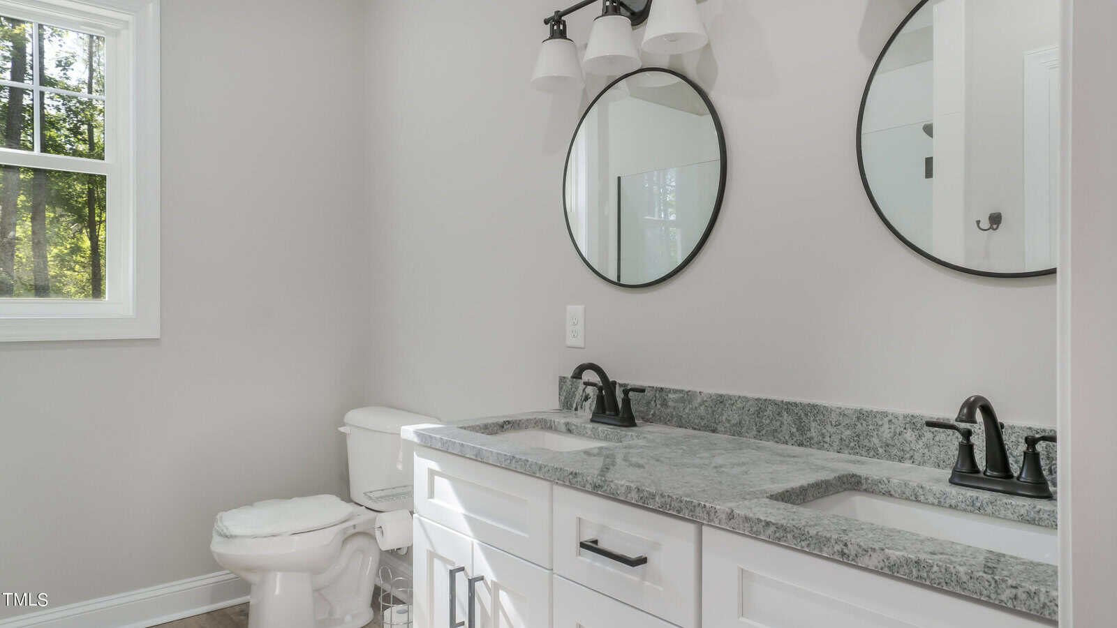 A modern bathroom with light gray walls features a double vanity with a gray marble countertop and two black-framed round mirrors above it. The vanity has white cabinets and black faucets. A white toilet is situated near a window that lets in natural light.