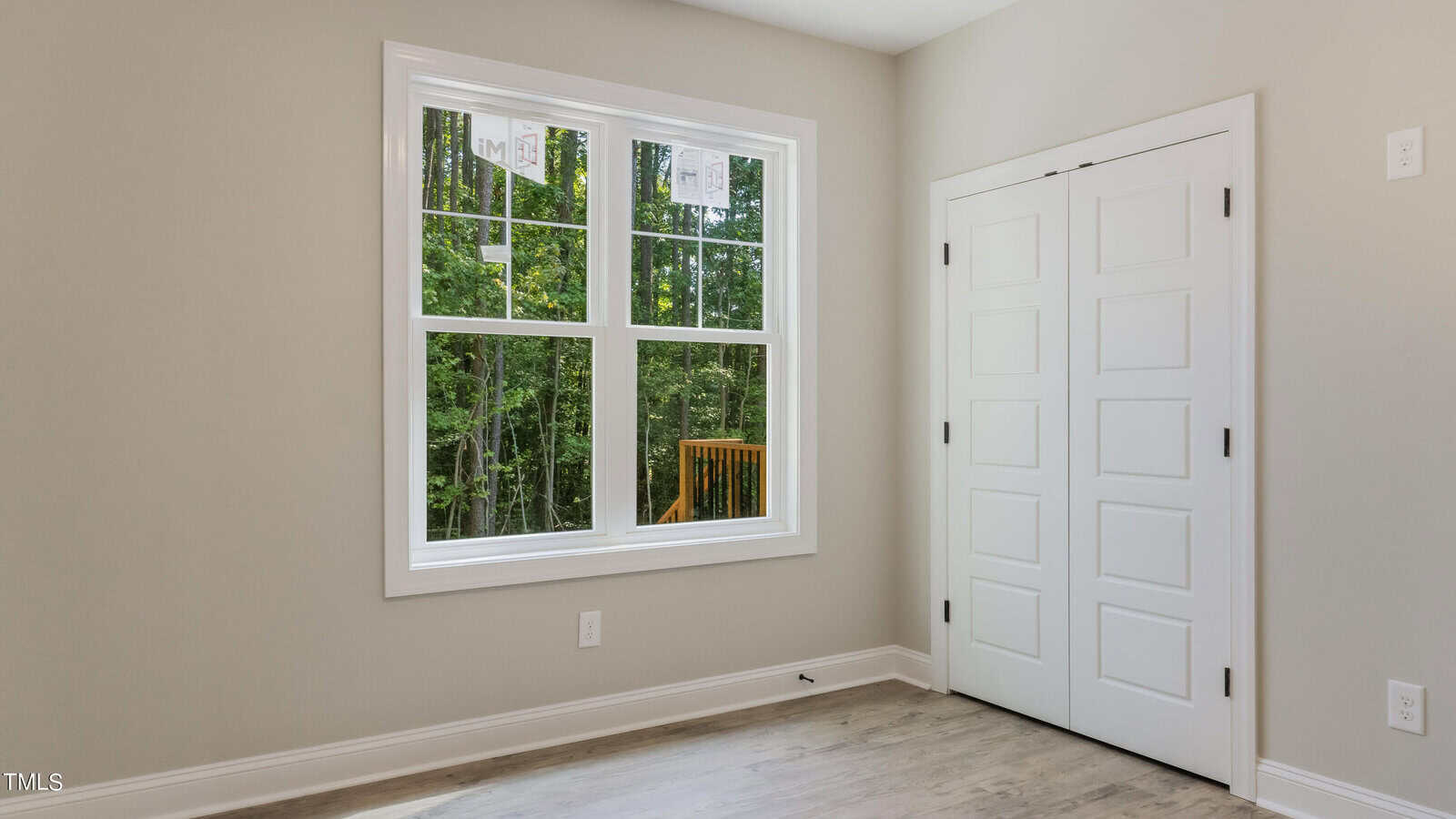 A bright, empty room with light-colored walls features a large window showing a view of trees outside. The floor is a light wood. Next to the window, there is a white double door closet. The room has a minimalist and clean design.