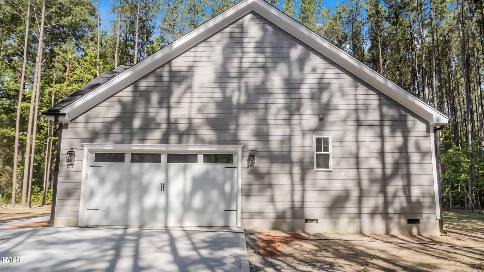 A side view of a modern, single-story house with gray horizontal siding and a white double garage door. A small window is next to the garage. Tall trees and their shadows are visible in the background, creating a serene, forested setting.