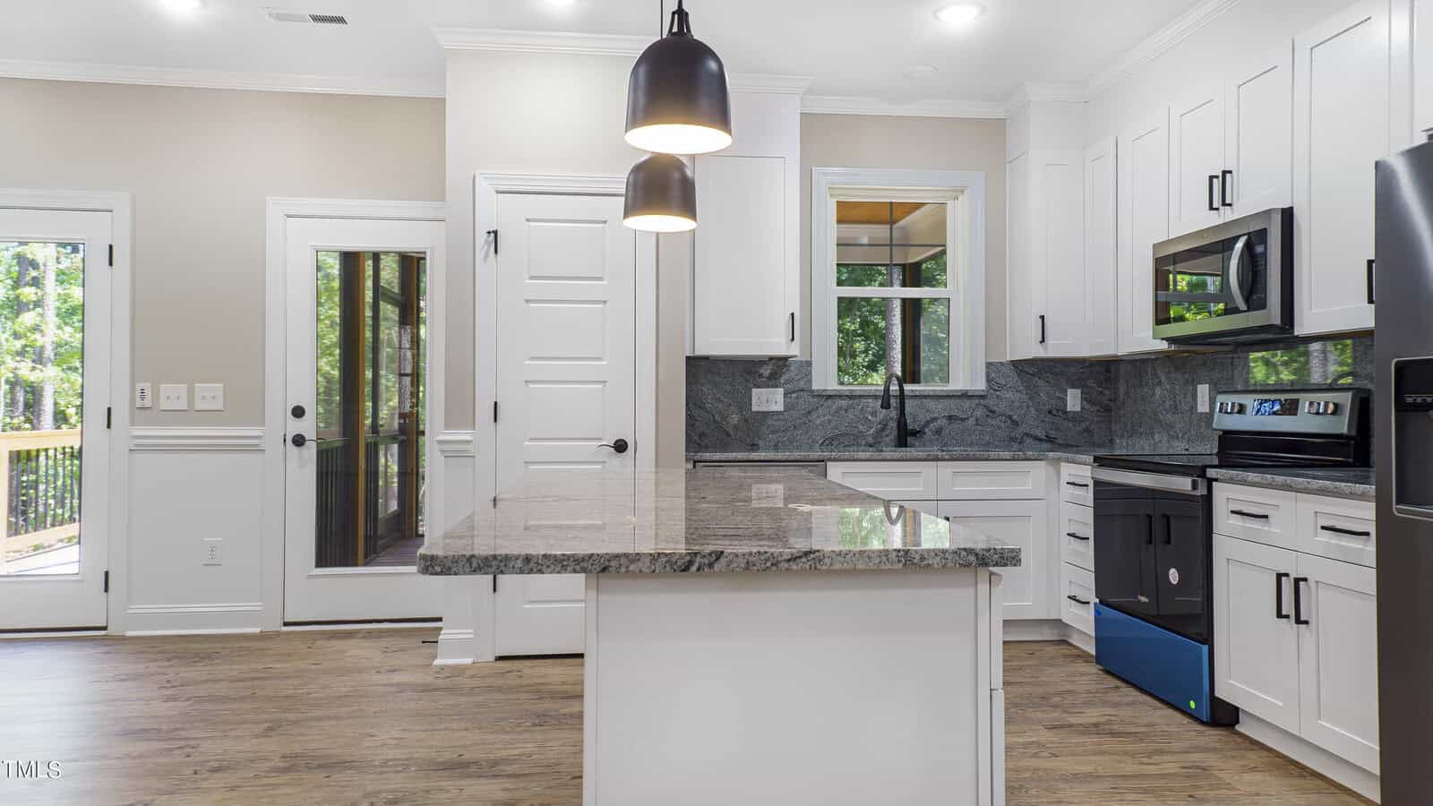 Modern kitchen with white cabinetry and black hardware. Includes a granite island countertop and stainless steel appliances, including a stove and microwave. Two black pendant lights hang over the island. There is a door and a window providing natural light.