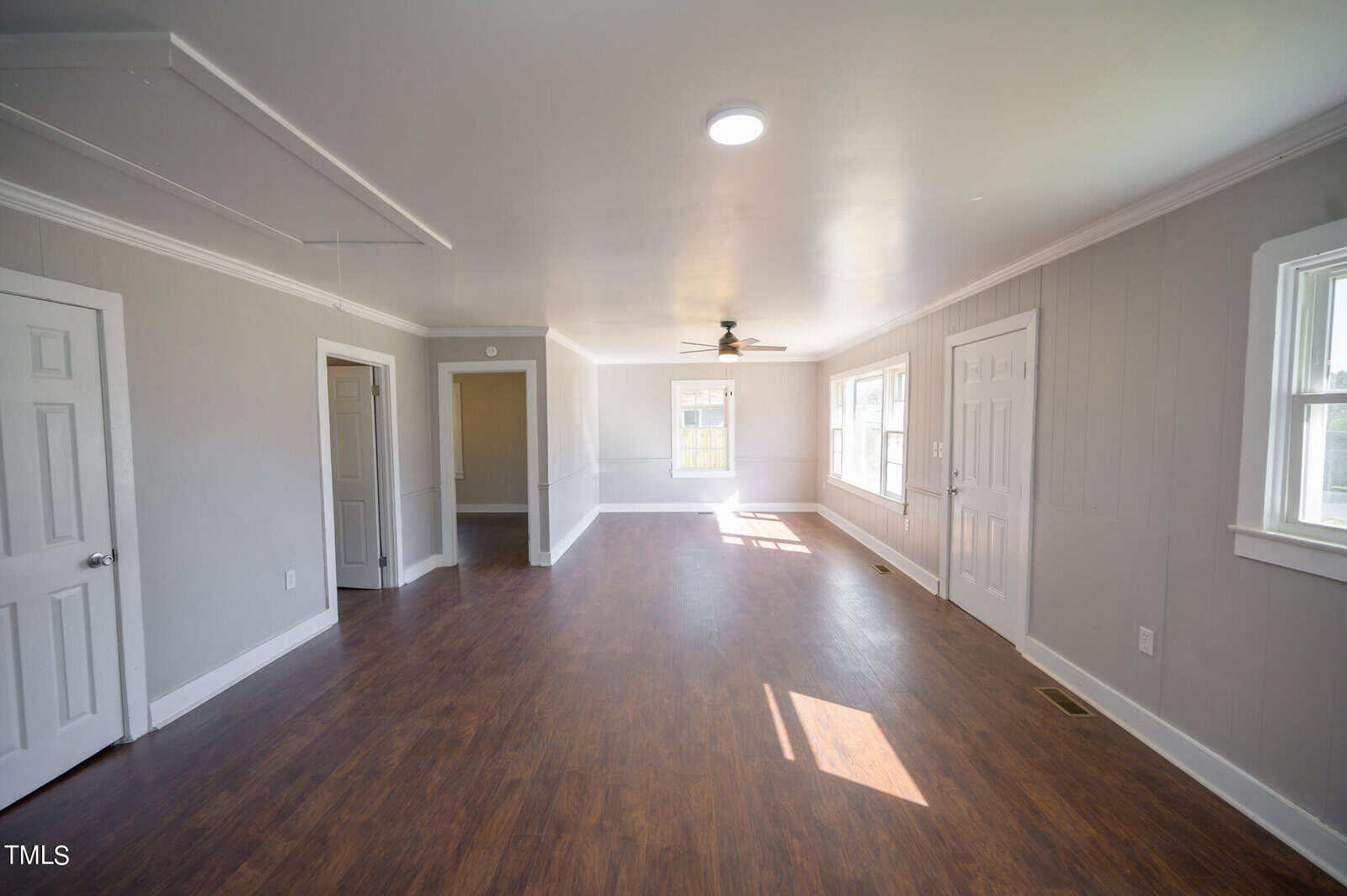 Spacious, empty room with hardwood floors, gray walls, and multiple doors. features ceiling fan, white trim, and natural light streaming in through windows.