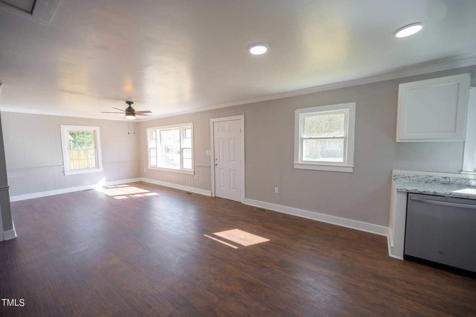 An empty, sunlit room with hardwood floors, white and gray walls, a ceiling fan, a door, two windows, and a kitchen counter with cabinets on the right.
