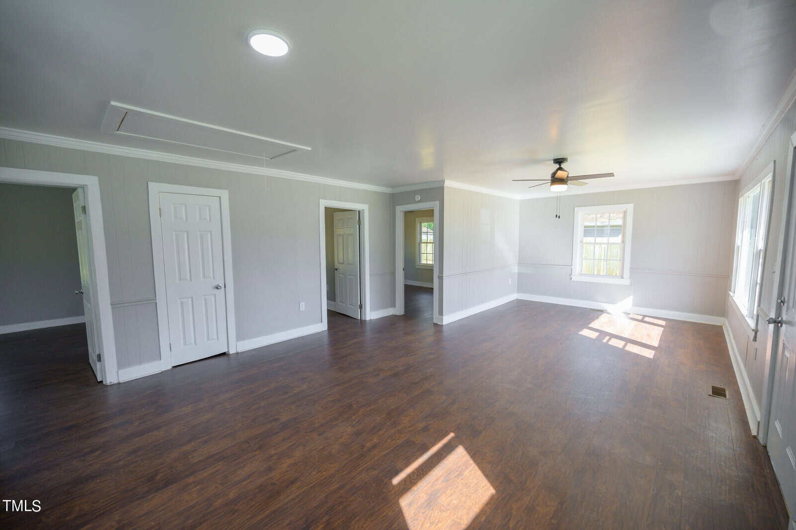 A spacious, empty room with polished wooden floors, white walls, and multiple doors. features include a ceiling fan, overhead lights, and a window letting in natural light.