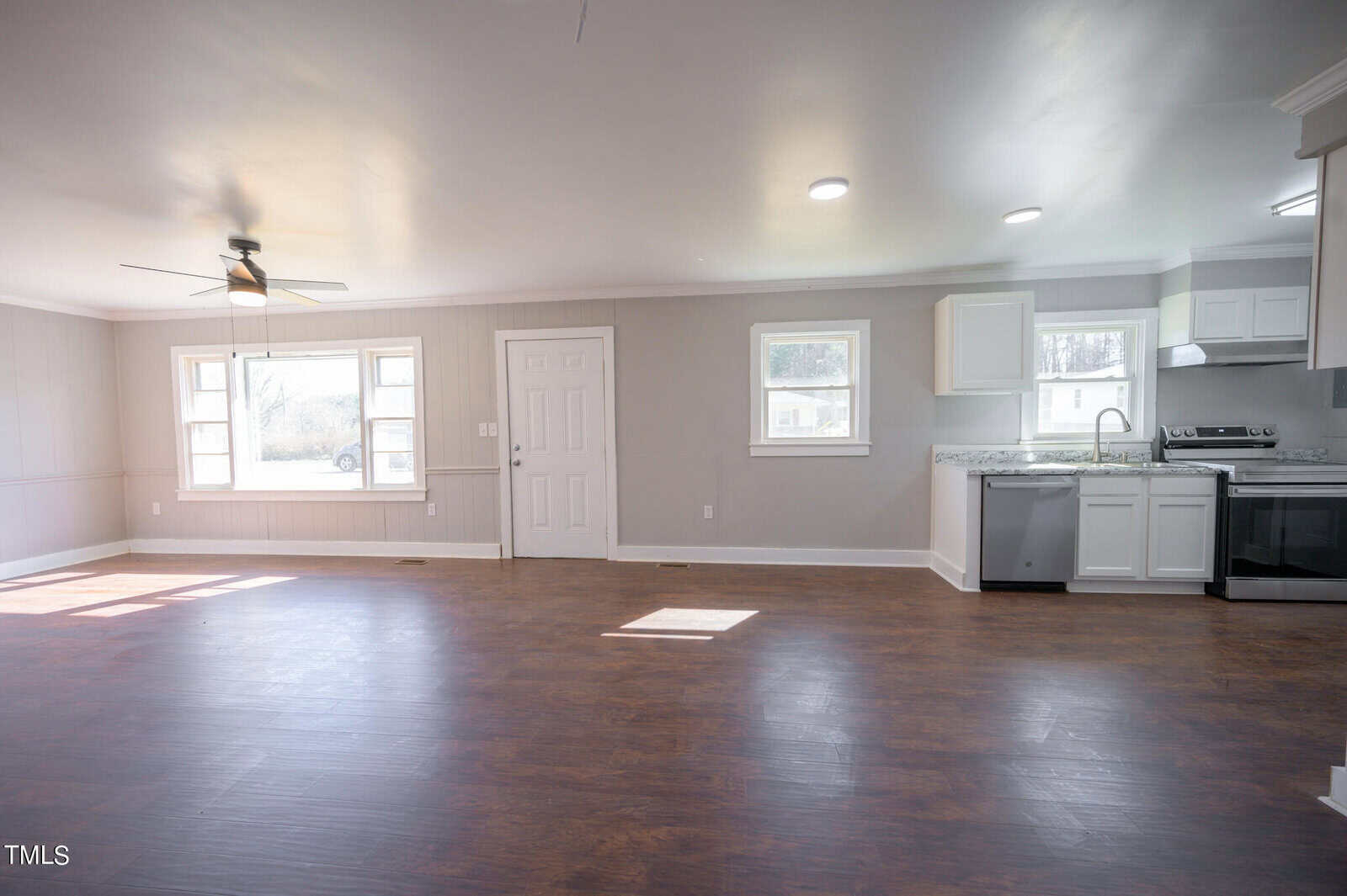 Spacious, well-lit room featuring dark hardwood floors, white walls, and a modern kitchen with white cabinets and granite countertops. there's a ceiling fan above and multiple windows providing natural light.