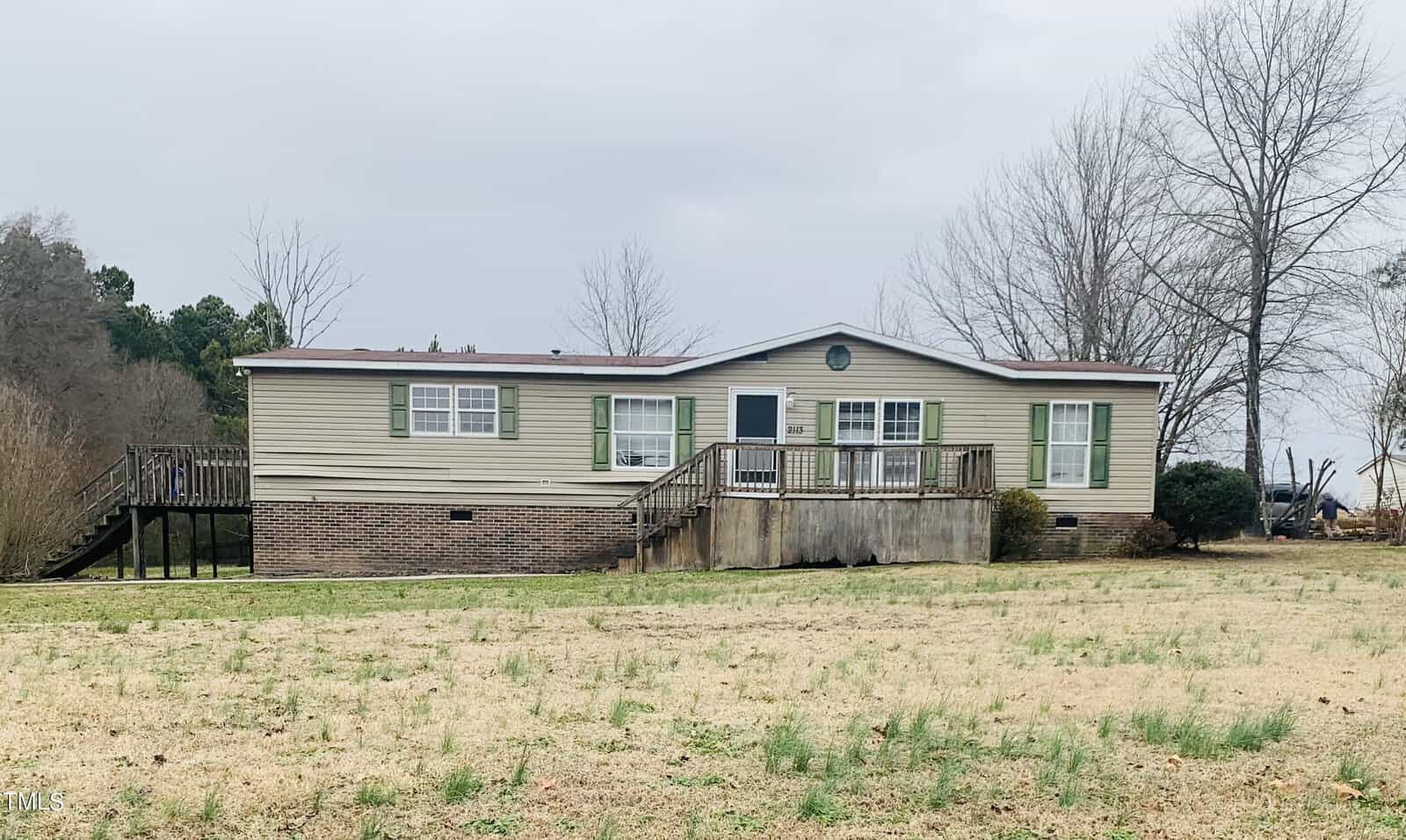 A single-story modular home located at 2113 Newton Dairy Road, Henderson NC, with a beige exterior and white trimming, featuring a front wooden deck and stairs, set on a spacious grassy lot