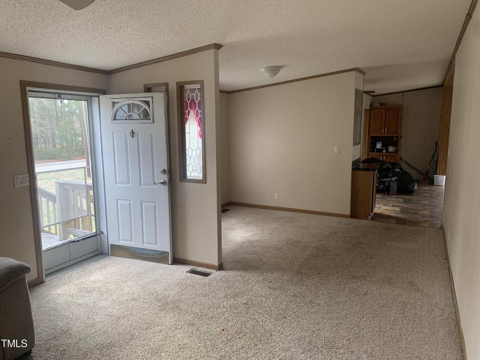 A view of an empty room with beige carpeting, featuring a front door with decorative glass and a side window, leading into a kitchen area with wooden cabinets.