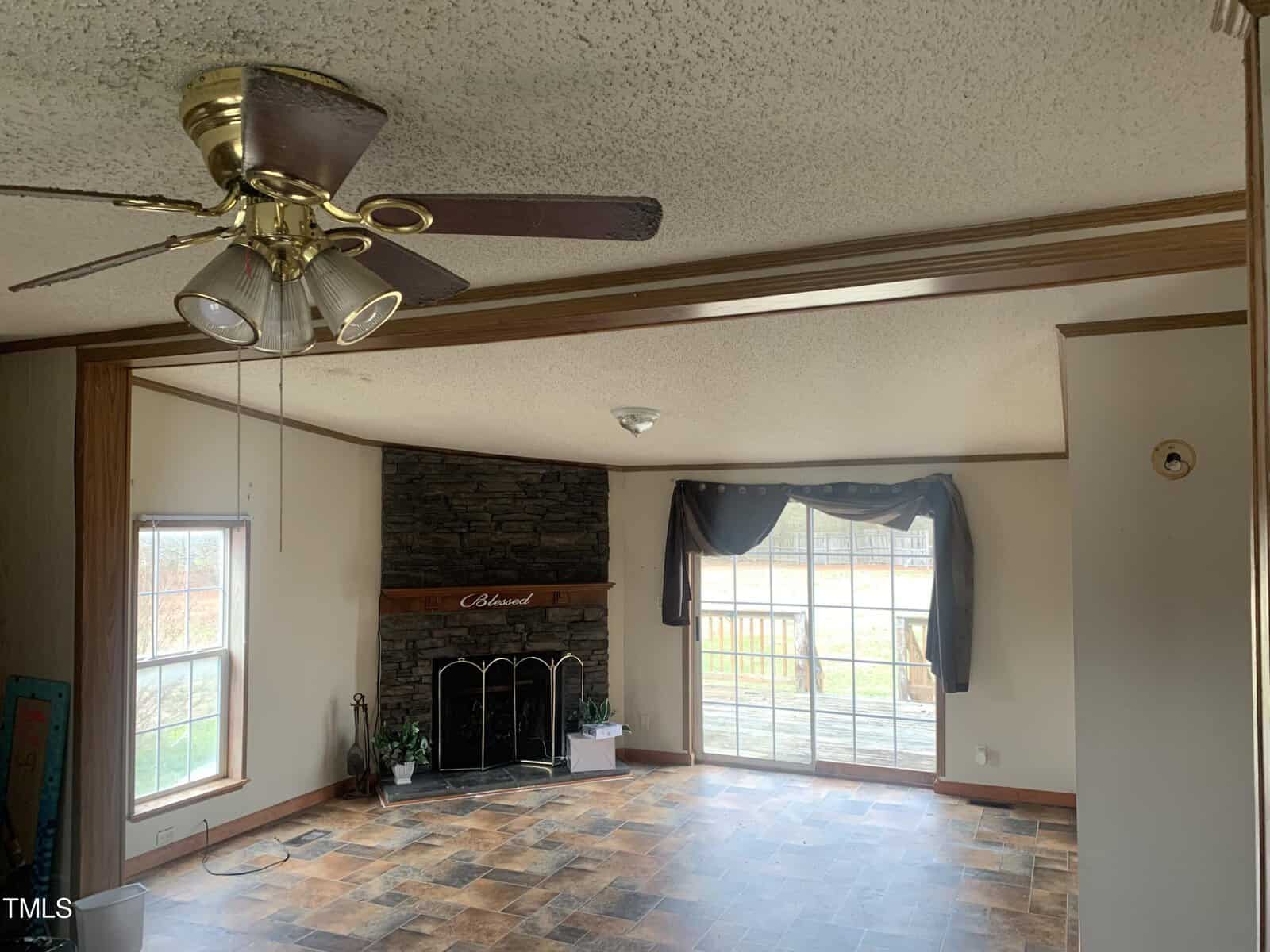 Interior of a room featuring a fireplace with a dark mantel, a ceiling fan above, and sliding glass doors with curtains leading to an outside area; the room has tiled flooring and wood-trimmed walls.