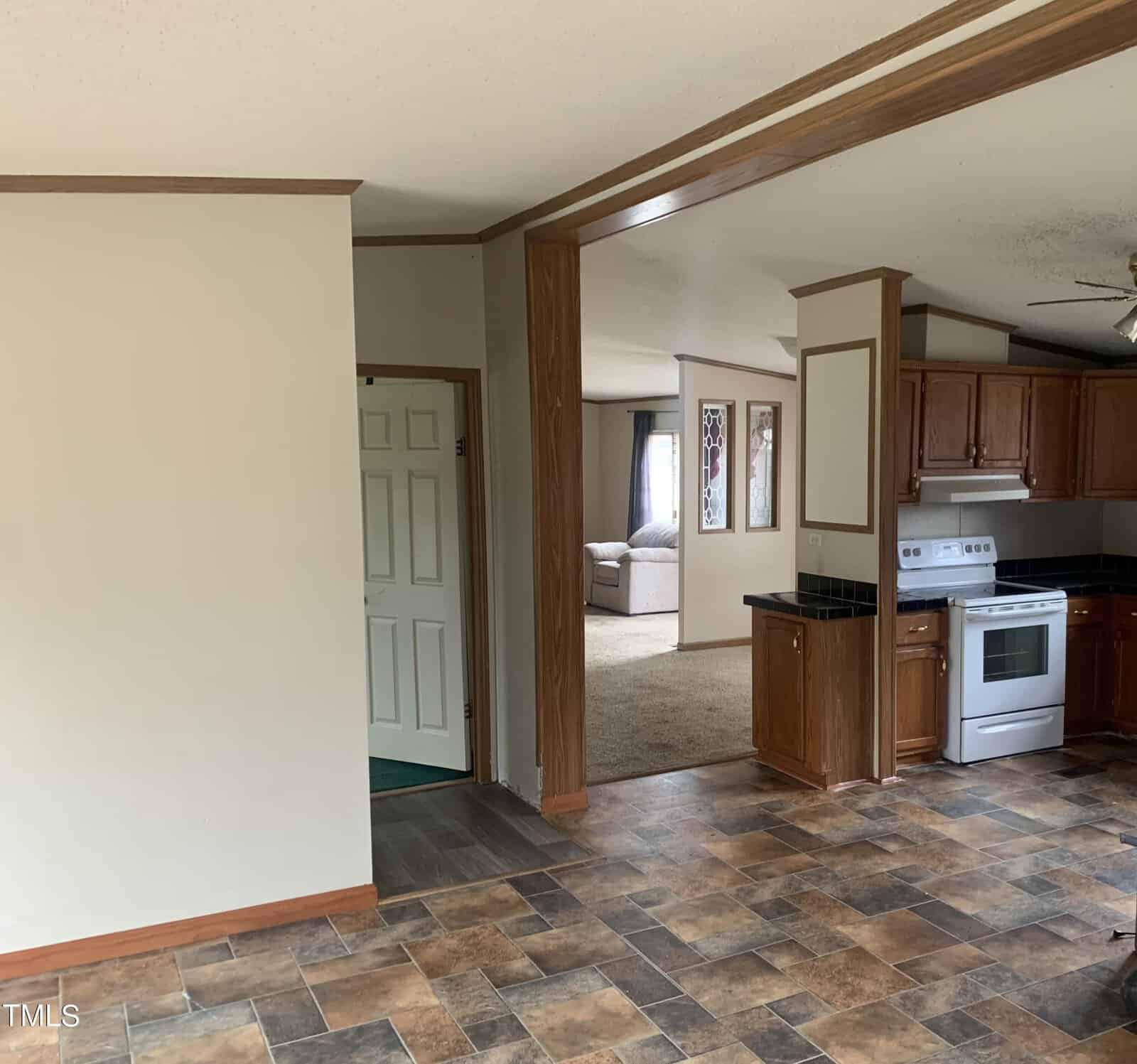 An interior view of a home showing a kitchen with wooden cabinets and white appliances adjacent to a living room area. the flooring transitions from tile in the kitchen to carpet in the living area.