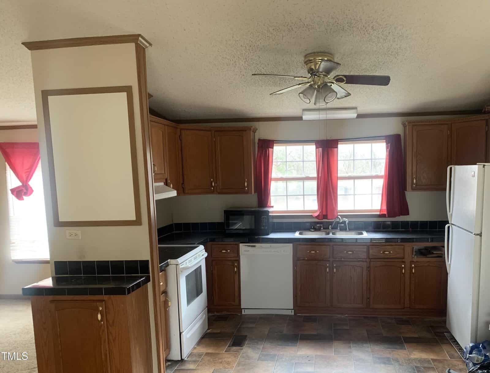 A kitchen interior with wooden cabinets, black countertops, a white refrigerator, and a stove, featuring red window curtains and a ceiling fan.