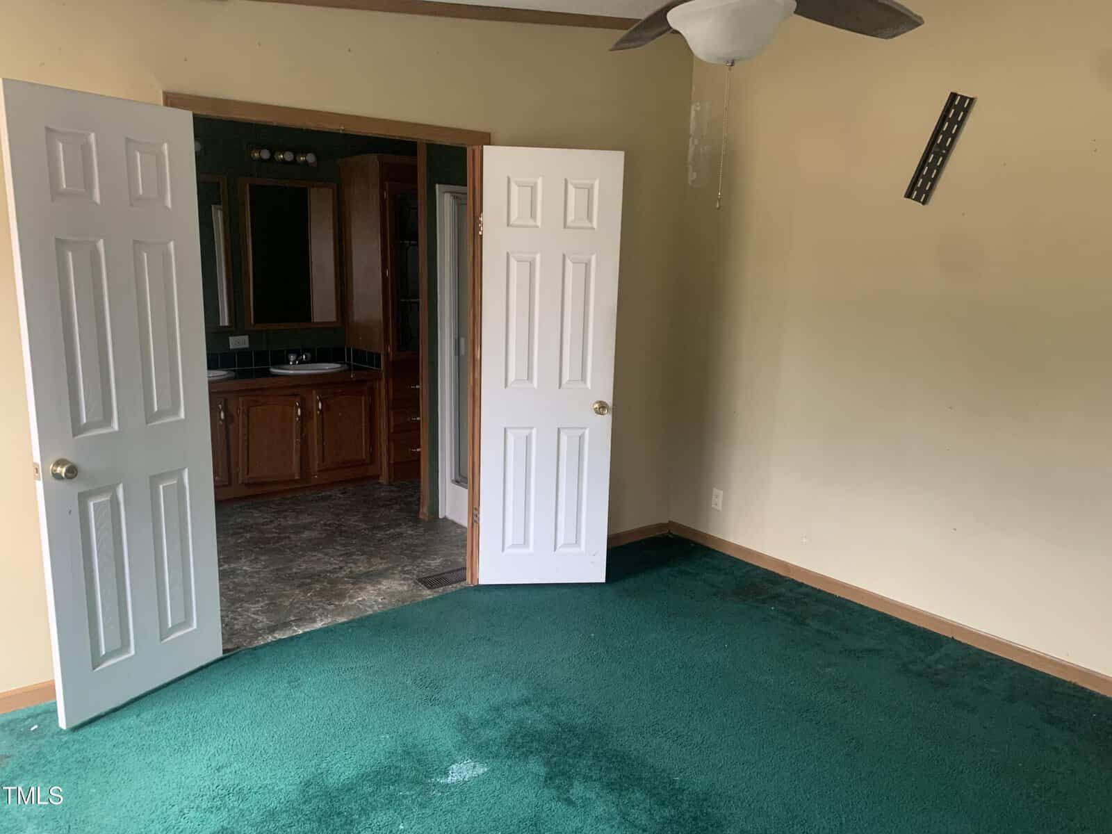 An empty room with green carpeting, tan walls, and an open white door on the left leading to a kitchen area with wooden cabinets. a ceiling fan is mounted above, and a black vent is on the right wall.