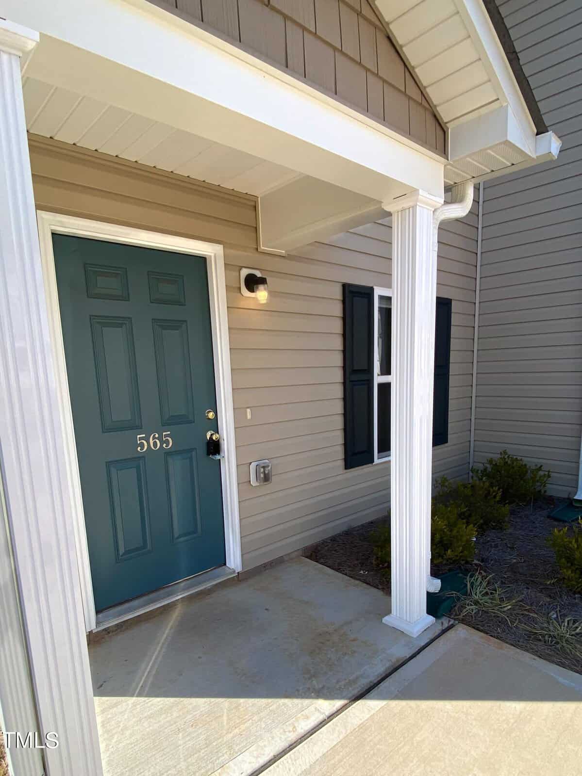 A front porch of a residential building featuring a teal door numbered 565 with a wall-mounted light fixture to the right, a window with black shutters, and a covered entrance with a white column.