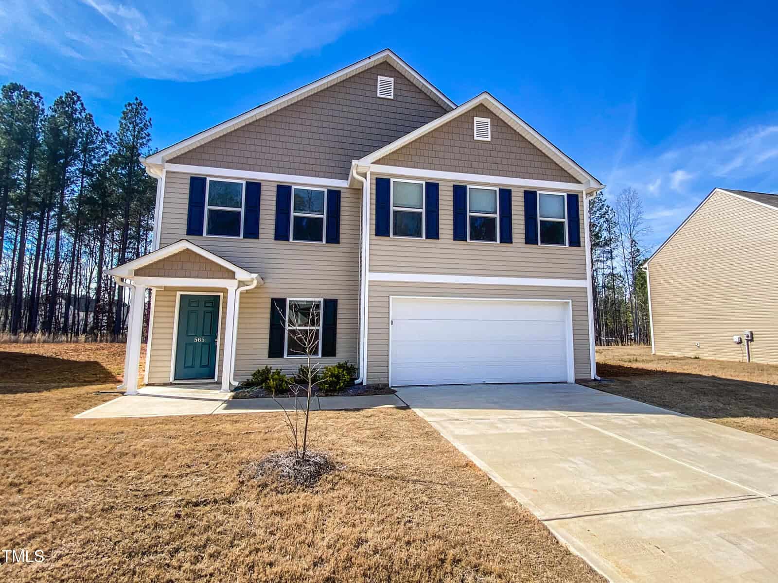 A newly constructed two-story house at 565 E Waycliff Road, Henderson NC, with a two-car garage and beige siding, featuring a turquoise front door under a clear blue sky.