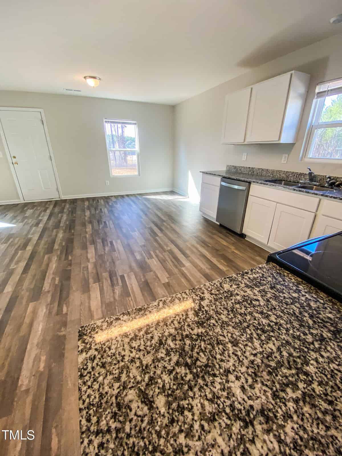 A modern kitchen with white cabinets, stainless steel appliances, and granite countertops, featuring a wood-laminated flooring that extends into an adjoining empty room lit by natural light from windows.