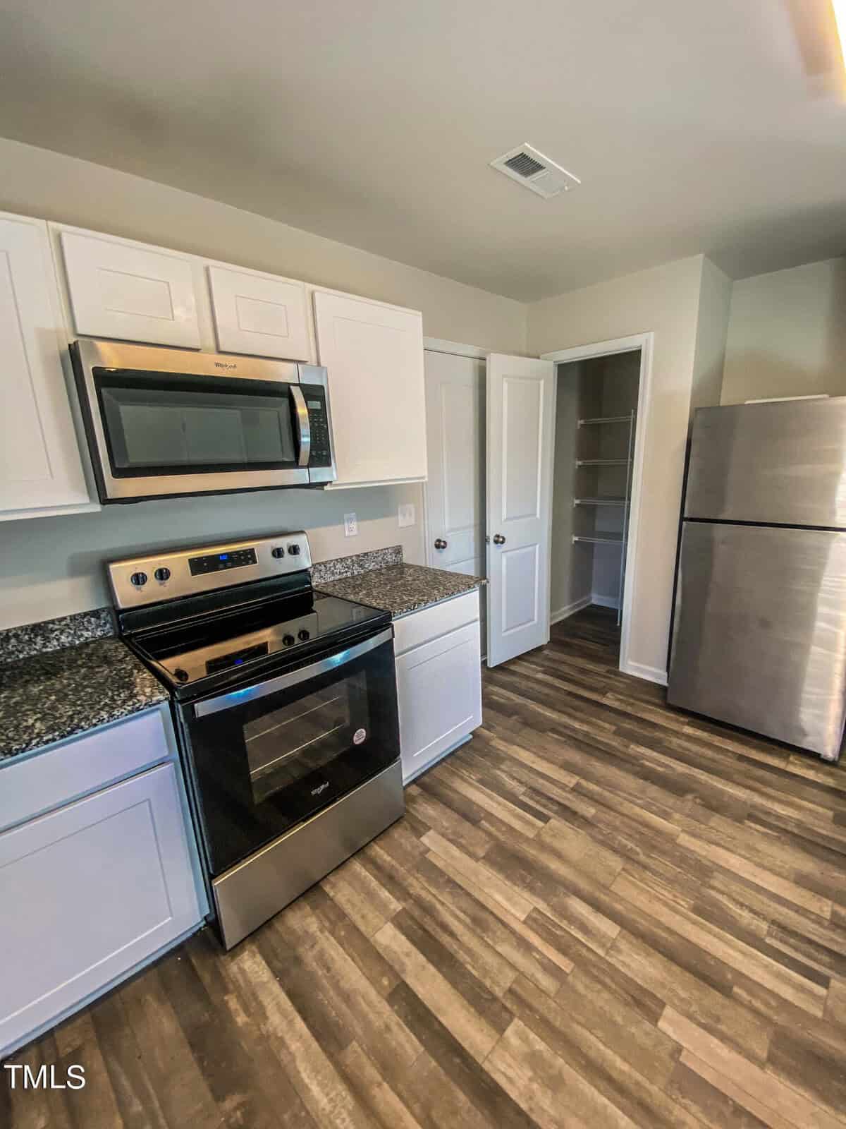 A modern kitchen equipped with stainless steel appliances, white cabinets, and a granite countertop, featuring a microwave, range oven, and a refrigerator, with hardwood-style flooring.