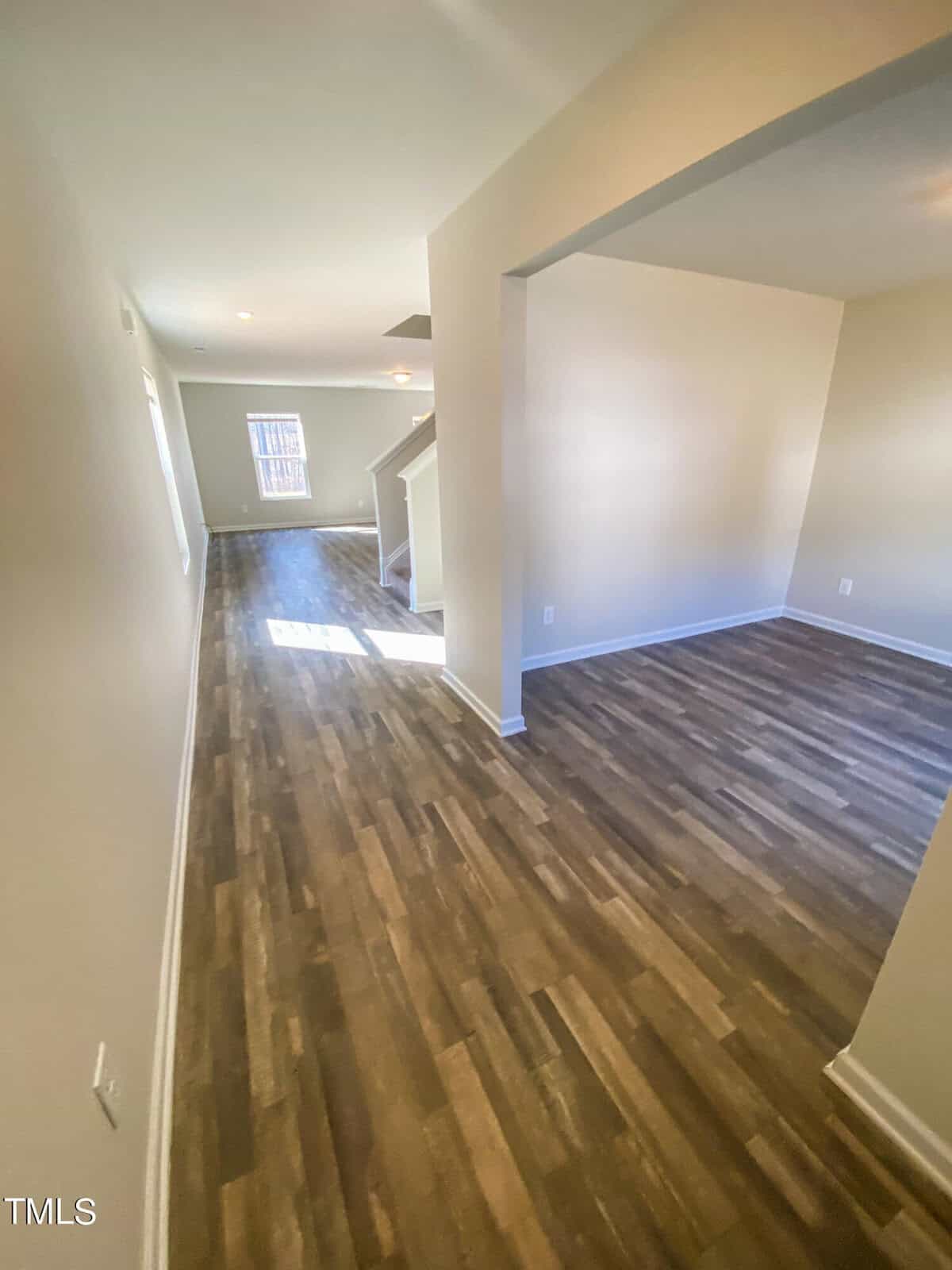 An empty room with hardwood floors, white walls, and an archway leading to an adjoining space. natural light enters through windows at the far end.