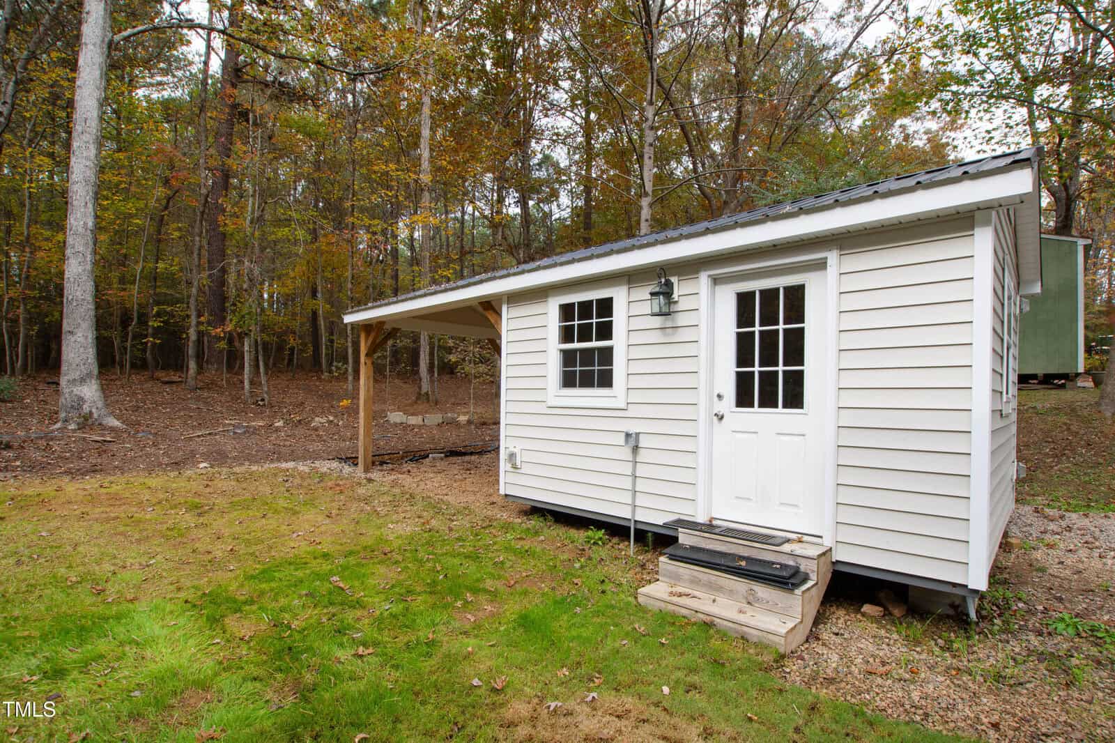 A small white shed with a covered side porch sits on a grassy area near a wooded forest. The shed has a white door, windows, and steps leading up to the entrance. Fallen leaves and trees surround the structure.