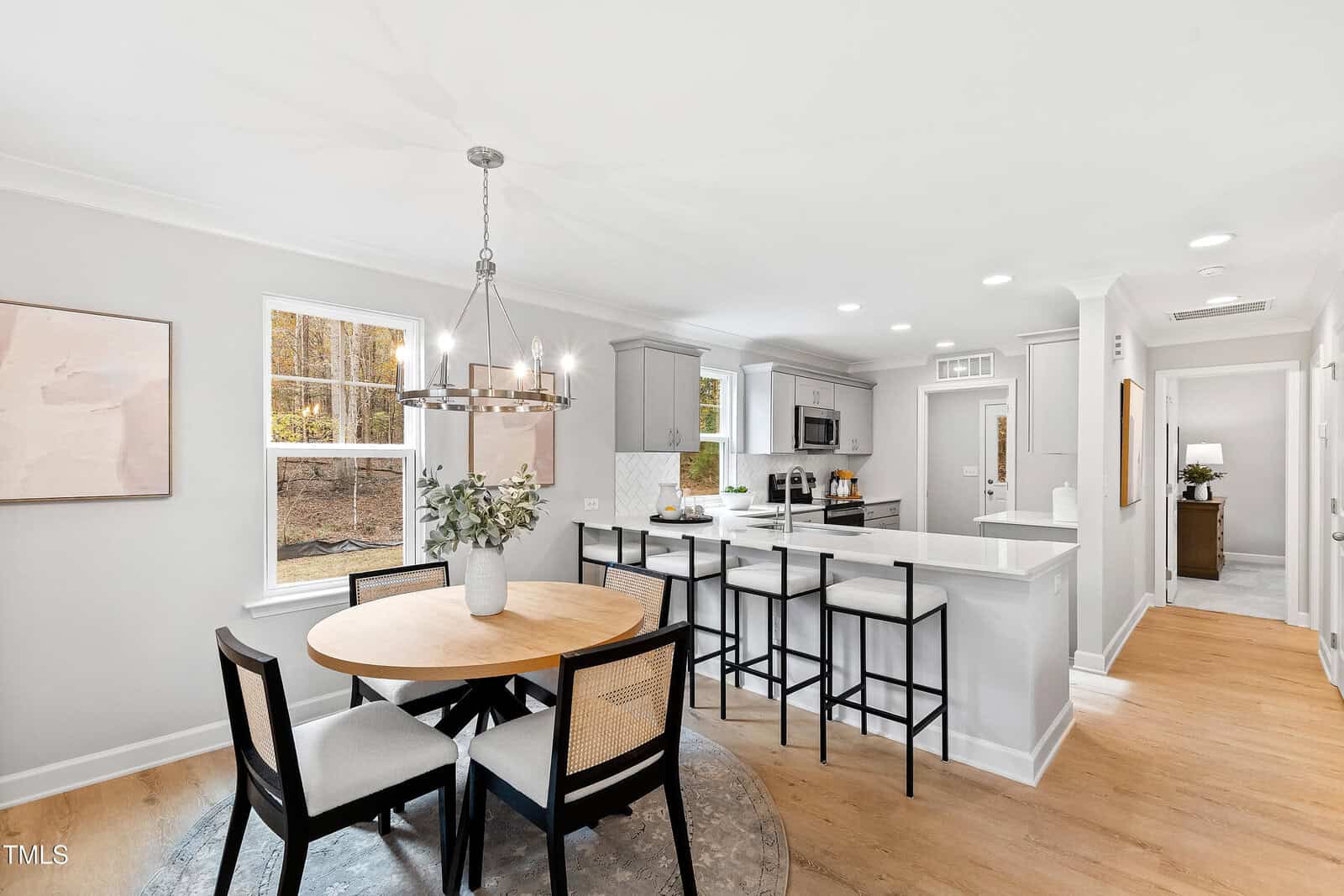 A modern kitchen and dining area with light gray cabinets, white countertops, a round wooden dining table with four chairs, and large windows letting in natural light. The space features neutral tones and contemporary decor.