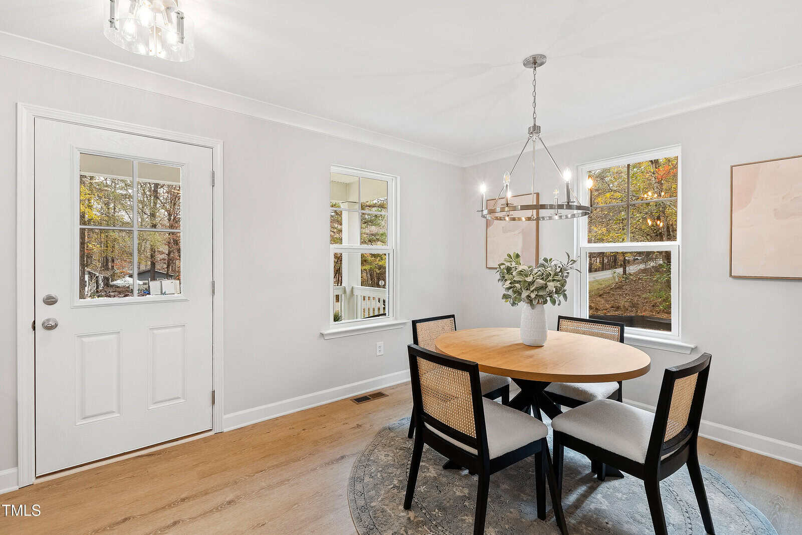 A bright dining area with light gray walls, wood flooring, and large windows. A round wooden table with four black chairs sits on a circular rug under a modern chandelier. A door with glass panes is on the left.