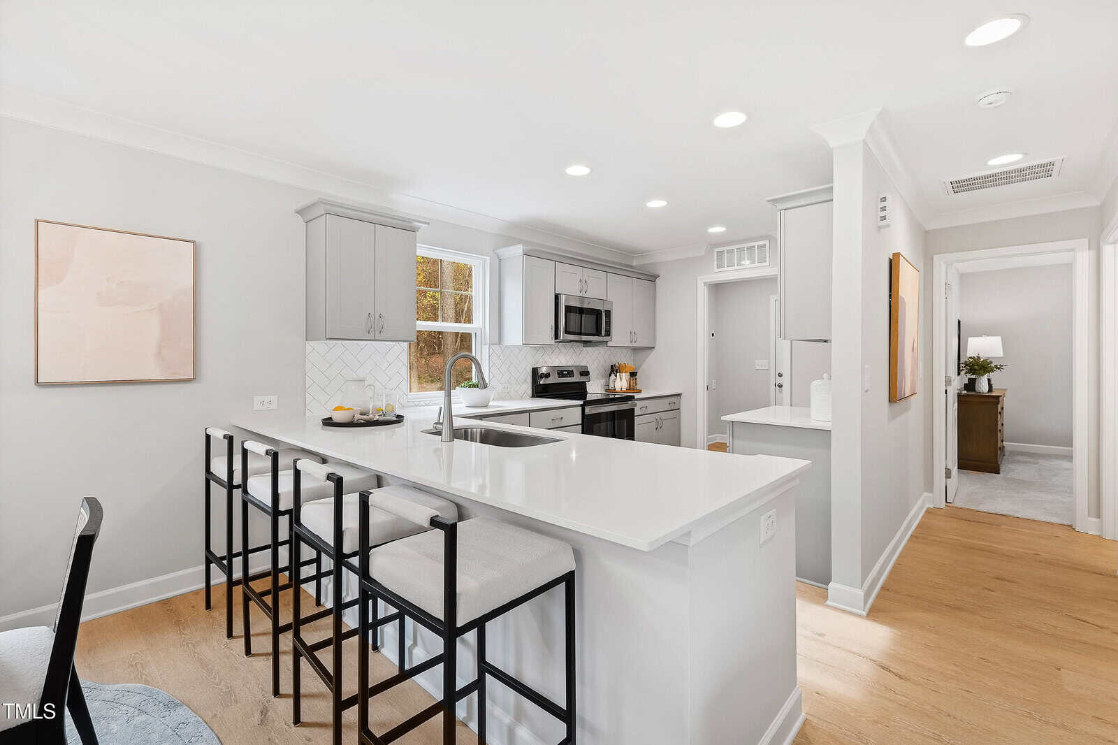 Bright, modern kitchen with white countertops, gray cabinets, stainless steel appliances, bar stools at an island, light wood flooring, and minimalist decor. A hallway in the background leads to other rooms.
