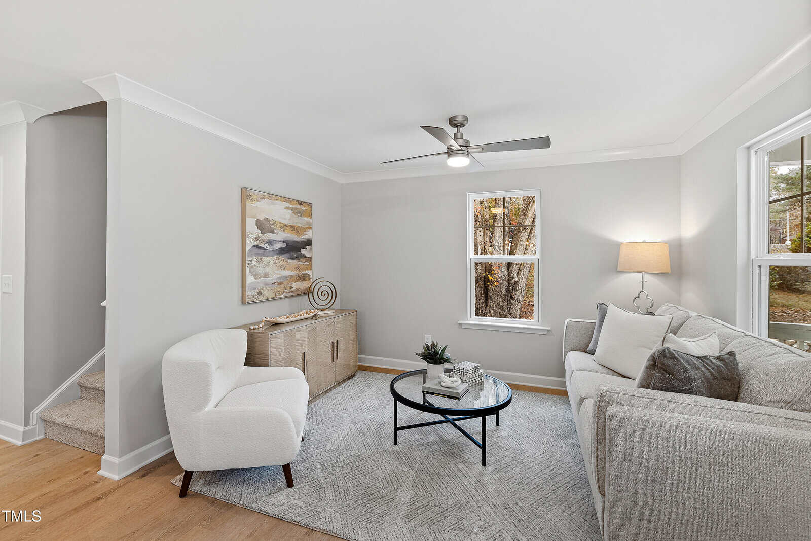Modern living room with light gray walls, a gray sofa, white armchair, round black coffee table, and neutral decor. A large window and lamp brighten the space. Carpeting and wood floors add contrast; stairs are visible on the left.