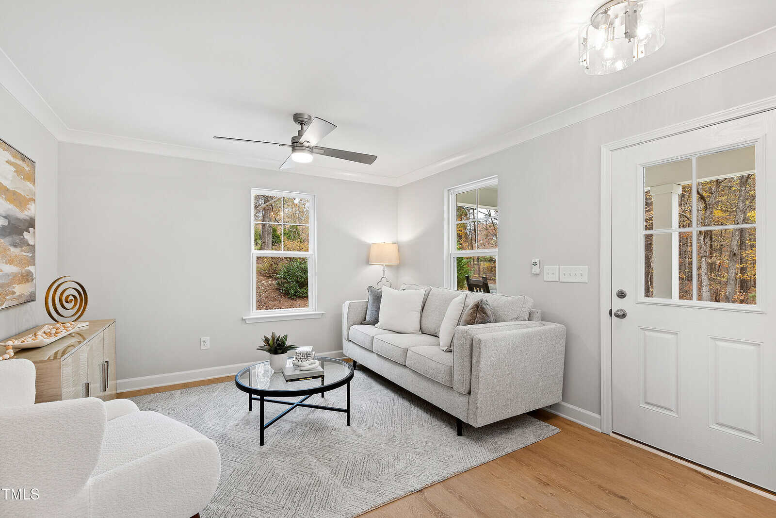 A bright living room with light-gray walls, a gray sofa, a white armchair, a round coffee table, a modern ceiling fan, and two windows letting in natural light. A door with glass panes is on the right.