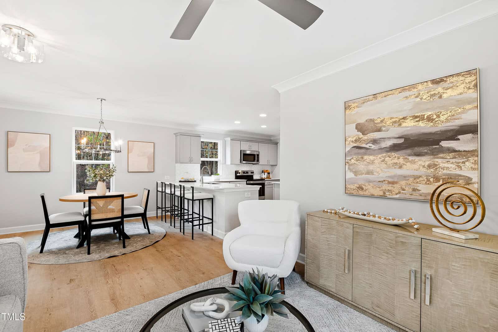 Modern open-concept living and dining area with neutral decor, round dining table with four chairs, kitchen with white cabinetry and bar stools, abstract art on the wall, and natural light from windows.