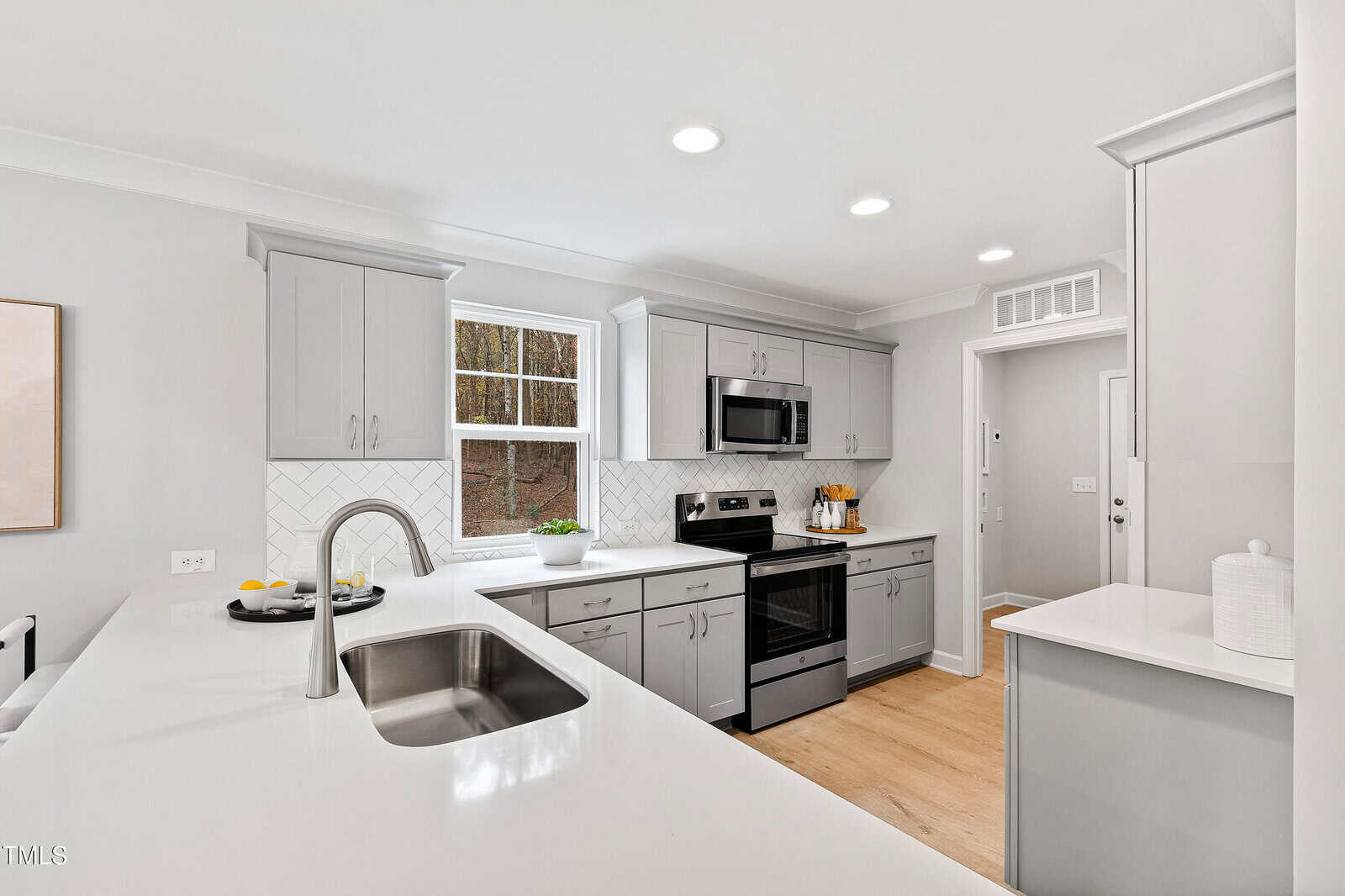 Modern kitchen with light gray cabinets, white countertops, stainless steel appliances, a large sink in a peninsula, and a window above the sink. The room has bright lighting and light wood flooring.